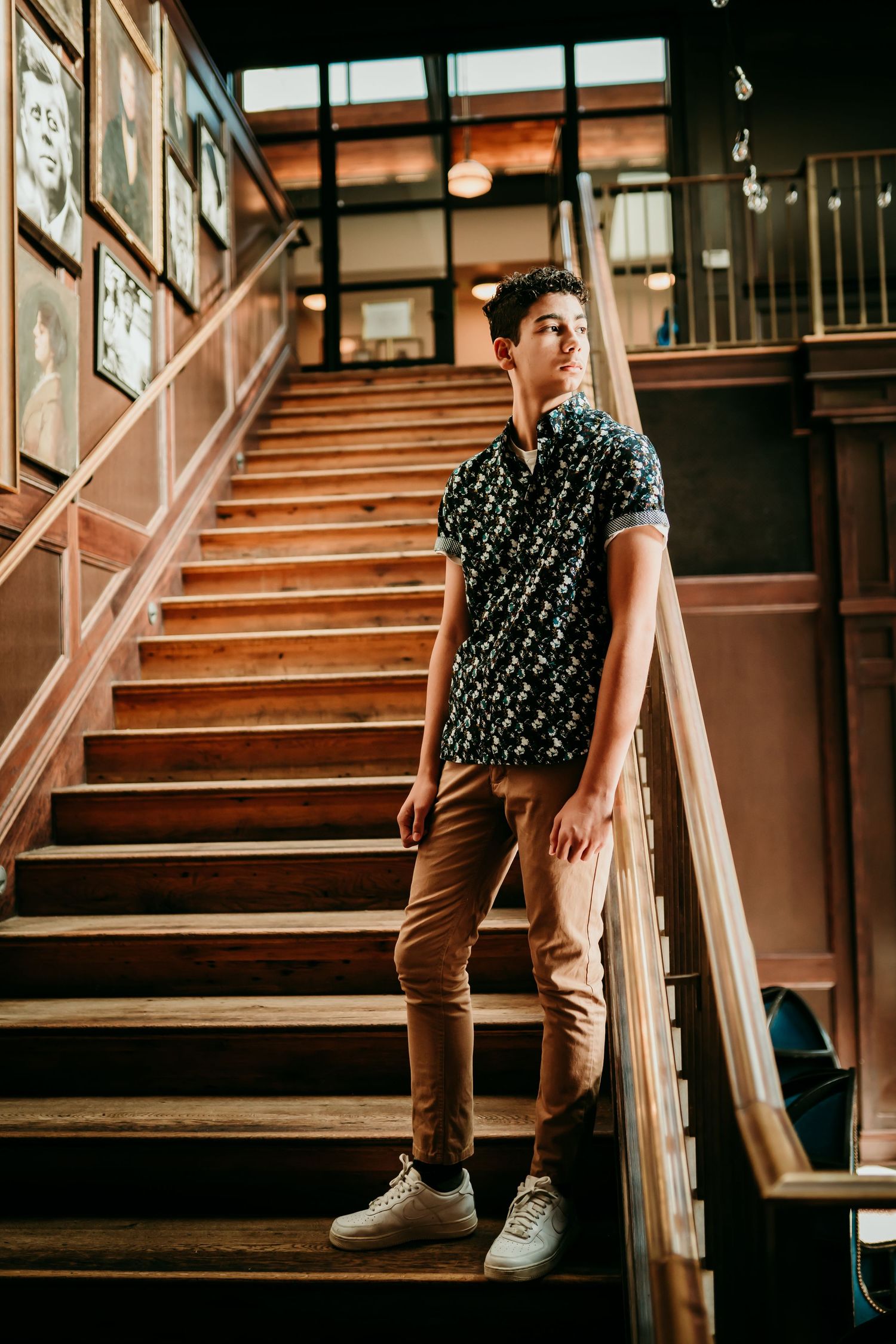 Young man in floral shirt and tan pants posing on wooden staircase in stylish indoor venue.