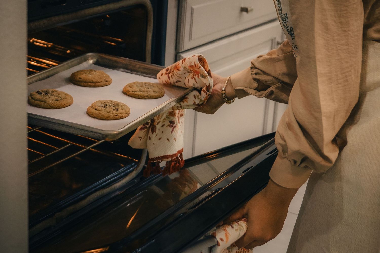 Person wearing oven mitts removing a tray of freshly baked chocolate chip cookies from a home oven.