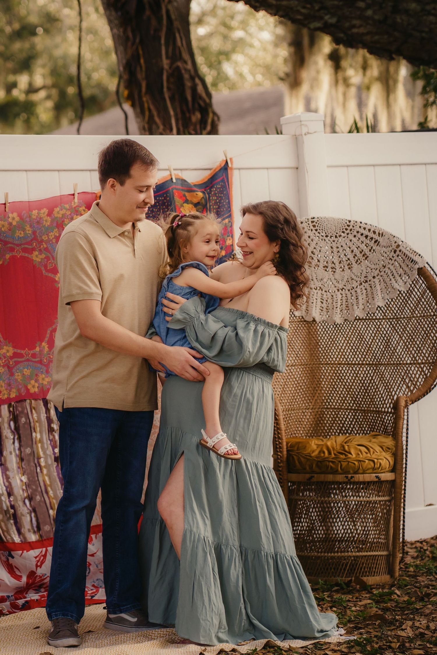 A happy family of three poses outdoors, parents holding their young daughter, with colorful fabric backdrop and wicker chair.