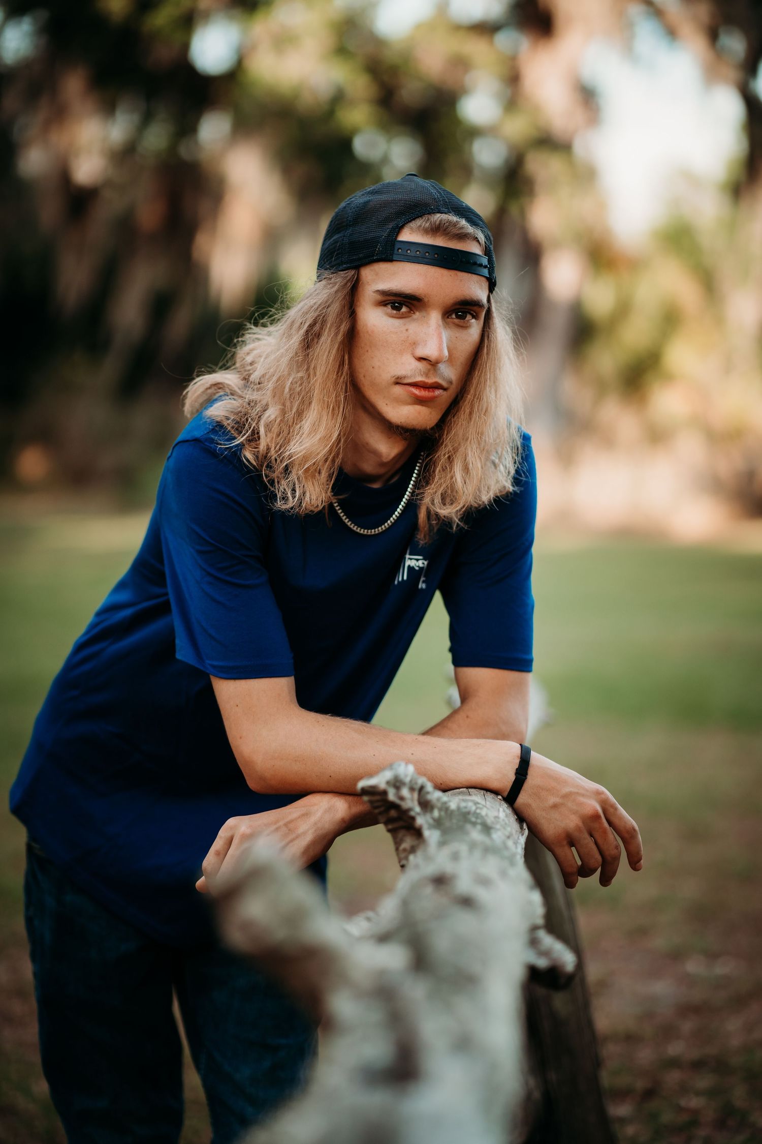 Young man with long blonde hair and backward cap leans on weathered log in outdoor park setting.