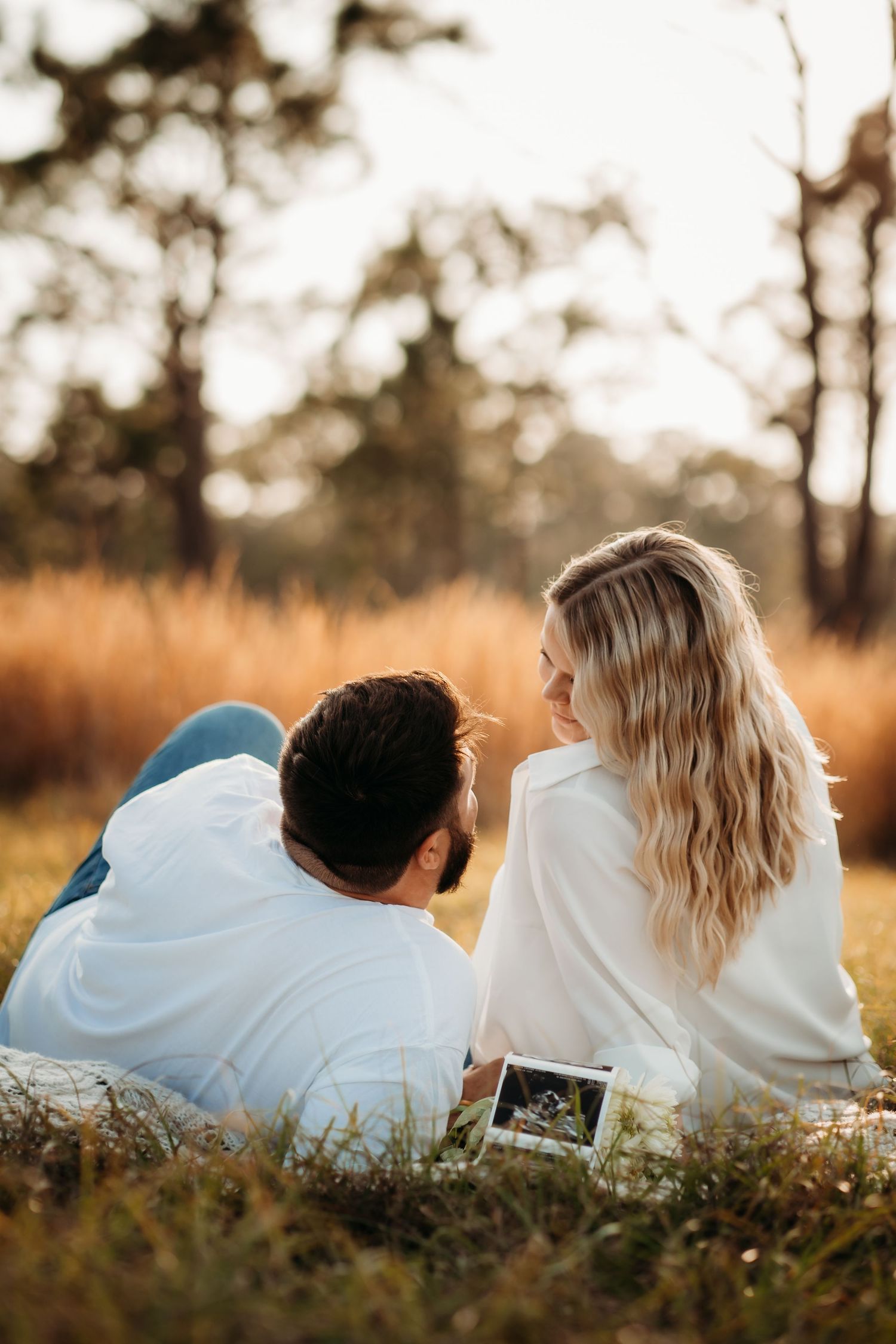 Couple lies on grass holding ultrasound photo in golden field at sunset, rear view.