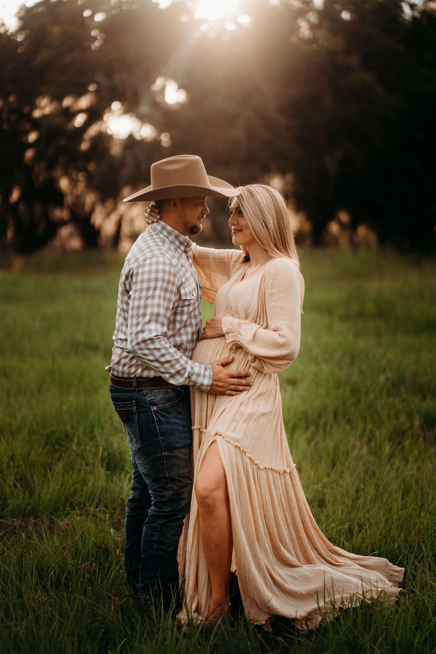 A couple embraces in a sunlit green field, the woman in a flowy beige maternity dress and man in a cowboy hat.