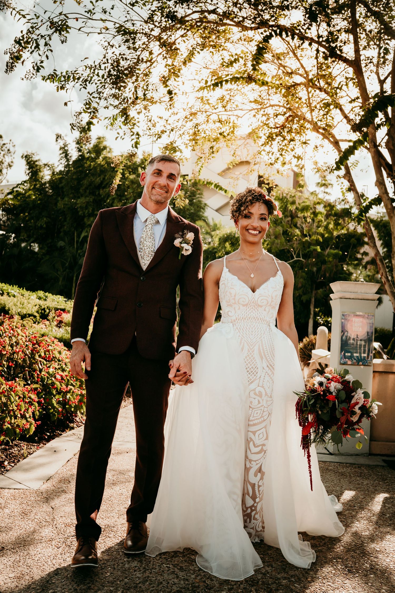 A smiling bride in a lace gown and groom in a dark suit hold hands outdoors at a garden wedding venue.
