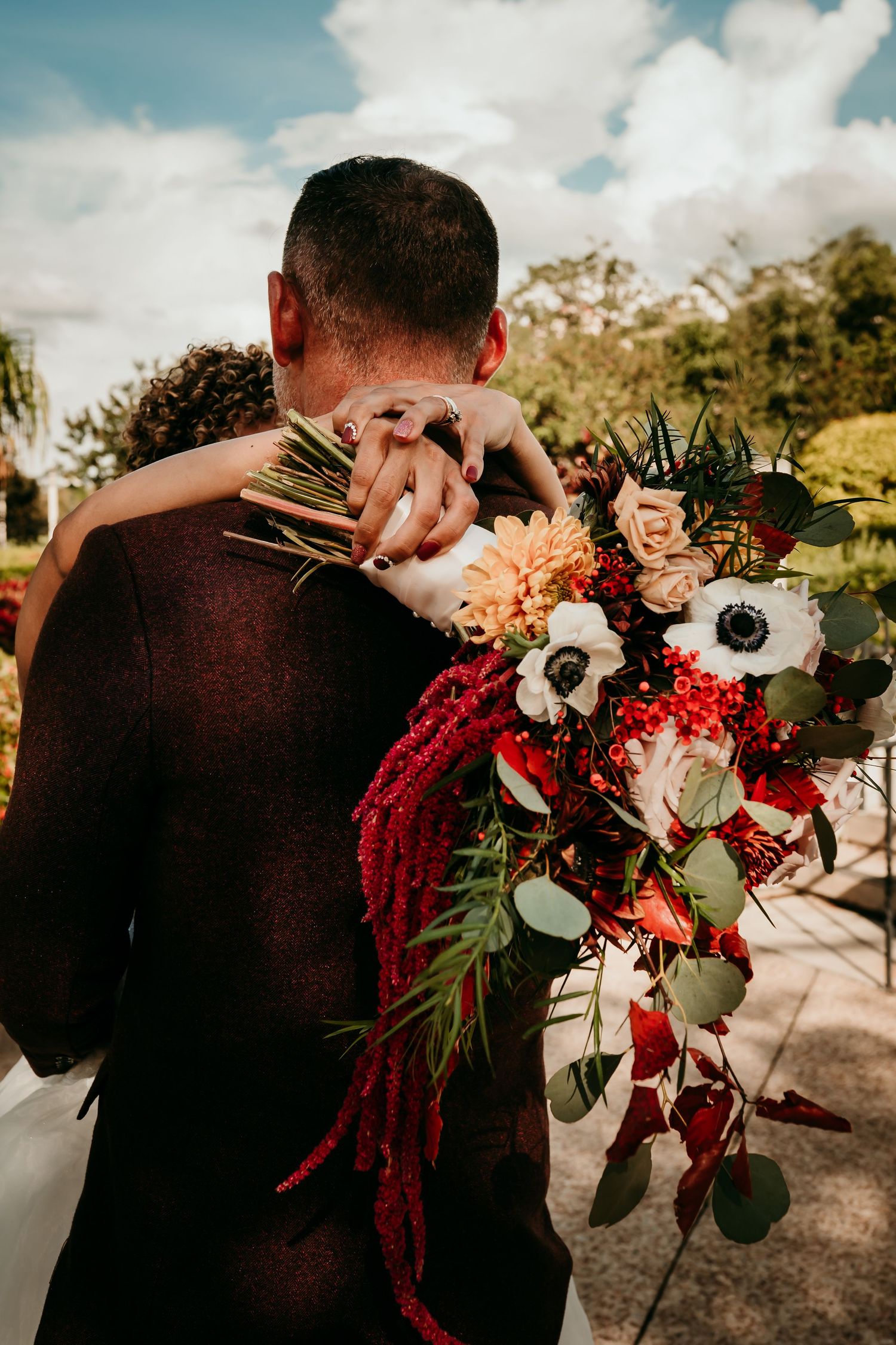 Bride holding colorful wedding bouquet with anemones and red amaranthus while embracing groom outdoors.