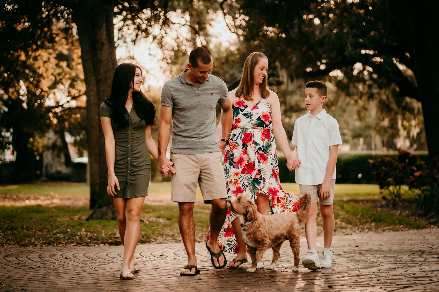 Family with dog walks along brick path under large oak trees at sunset.