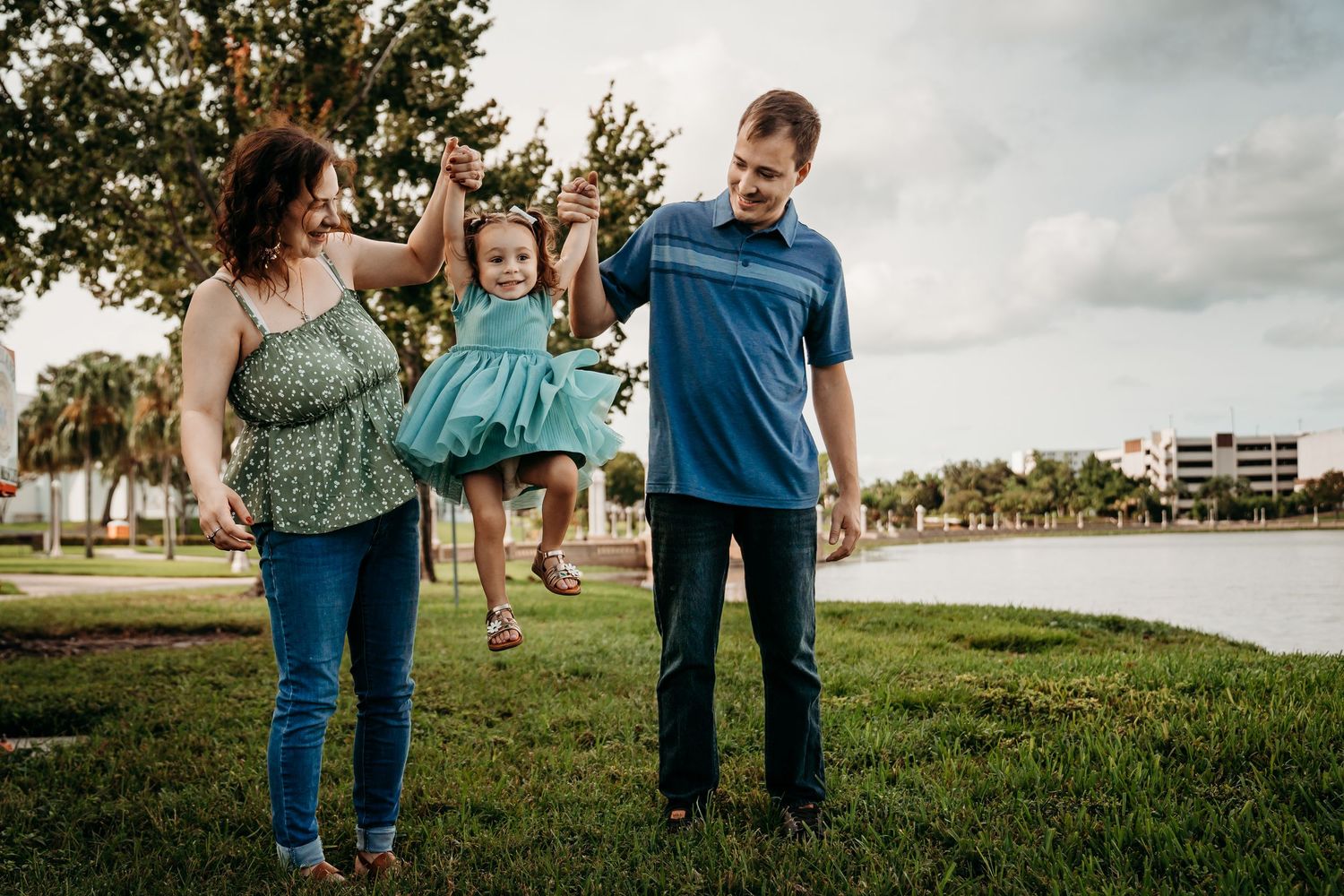 Parents swinging their laughing toddler daughter between them on a grassy lakeside park.