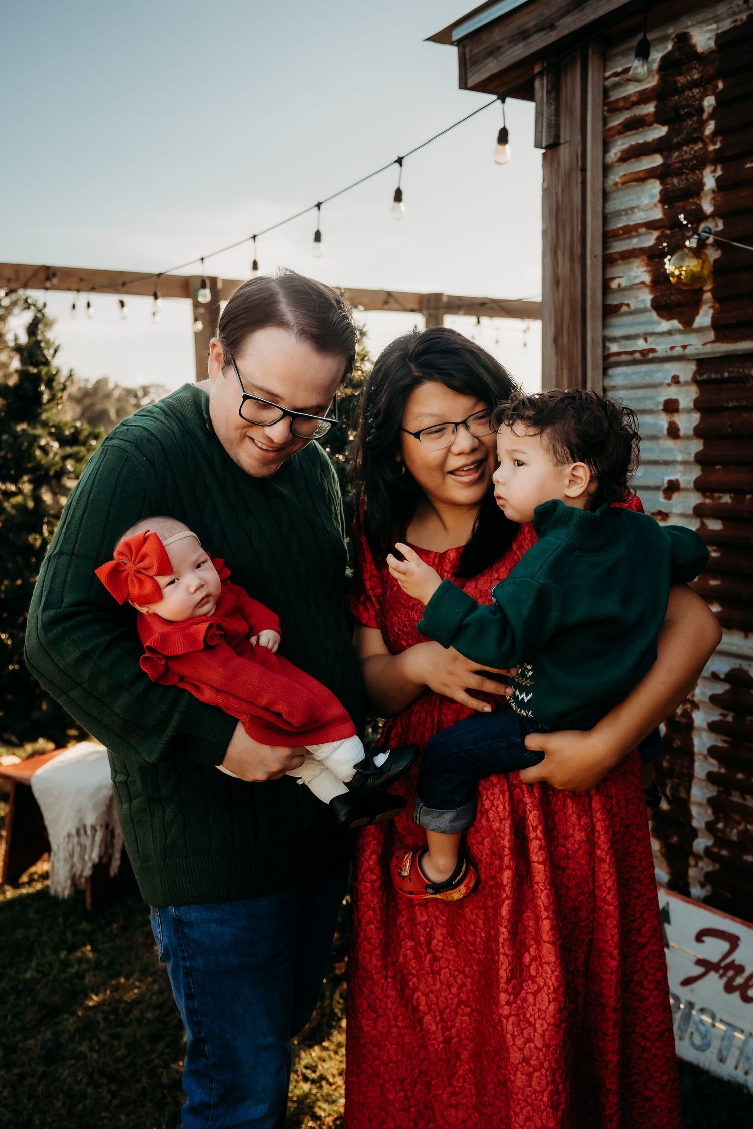 Young family of four in red and green holiday outfits admire their newborn at a Christmas tree farm.