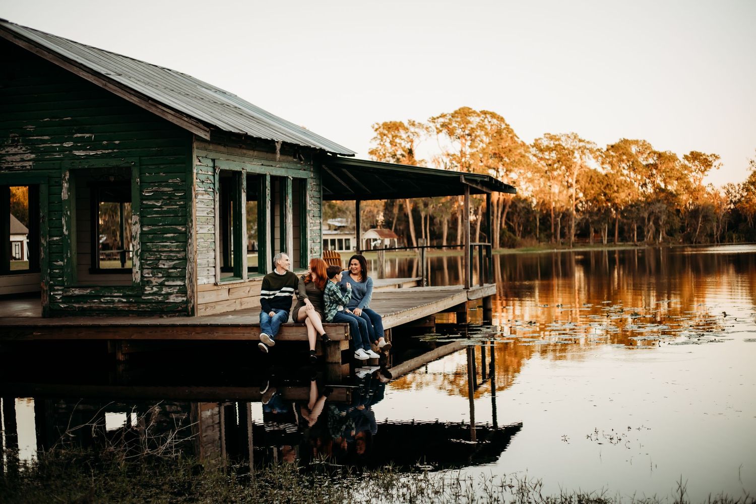 Couple sitting on dock beside rustic green boathouse at dusk.
