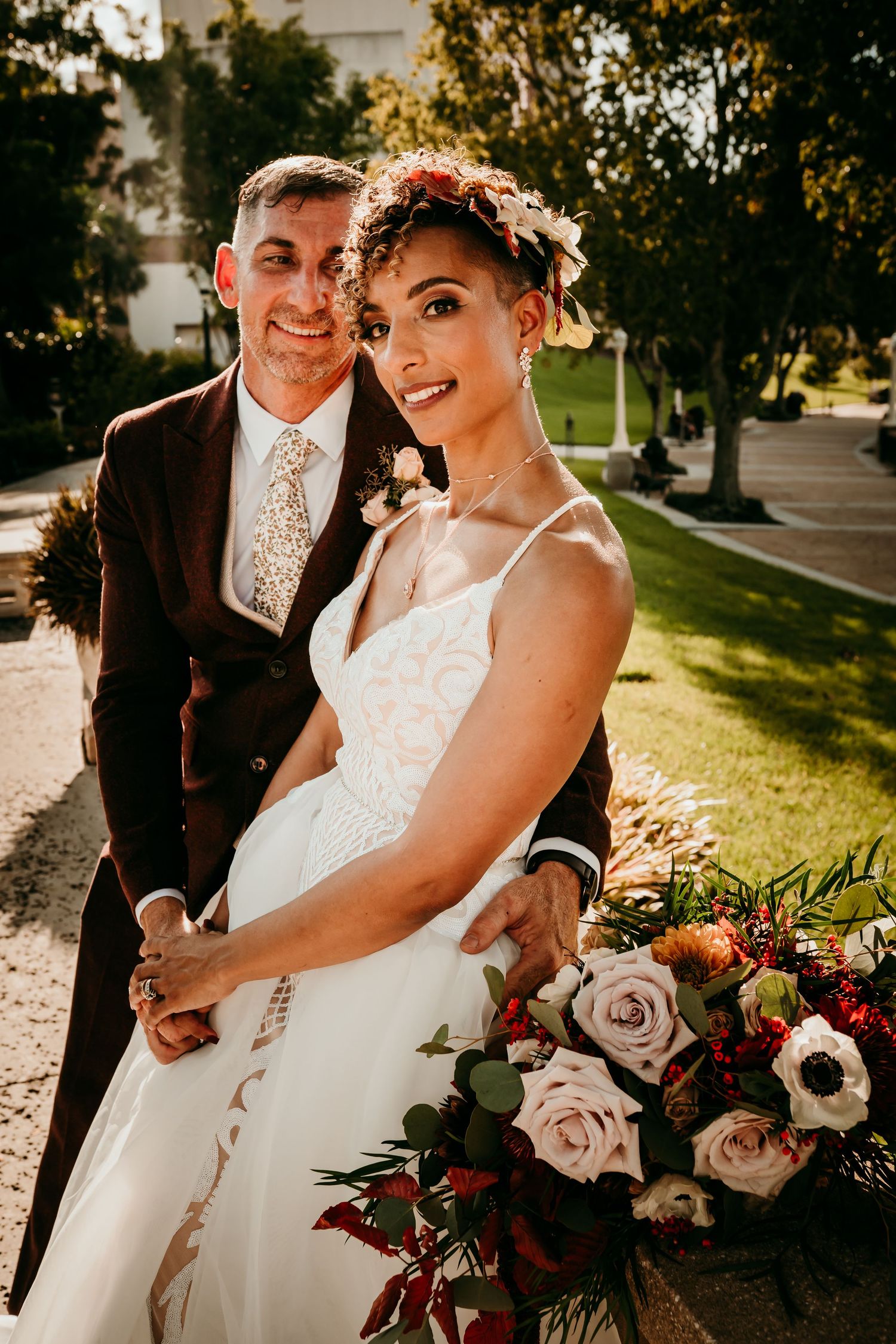 Bride in white lace gown with floral crown holds bouquet beside groom in brown suit outdoors.
