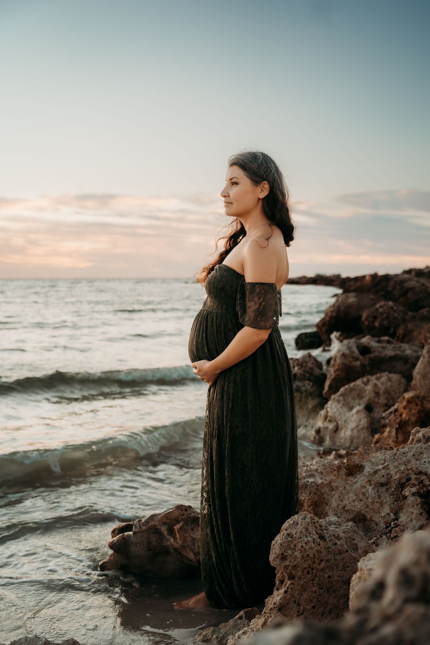 A pregnant woman in a dark off-shoulder gown stands on rocky shoreline at sunset, cradling her baby bump.