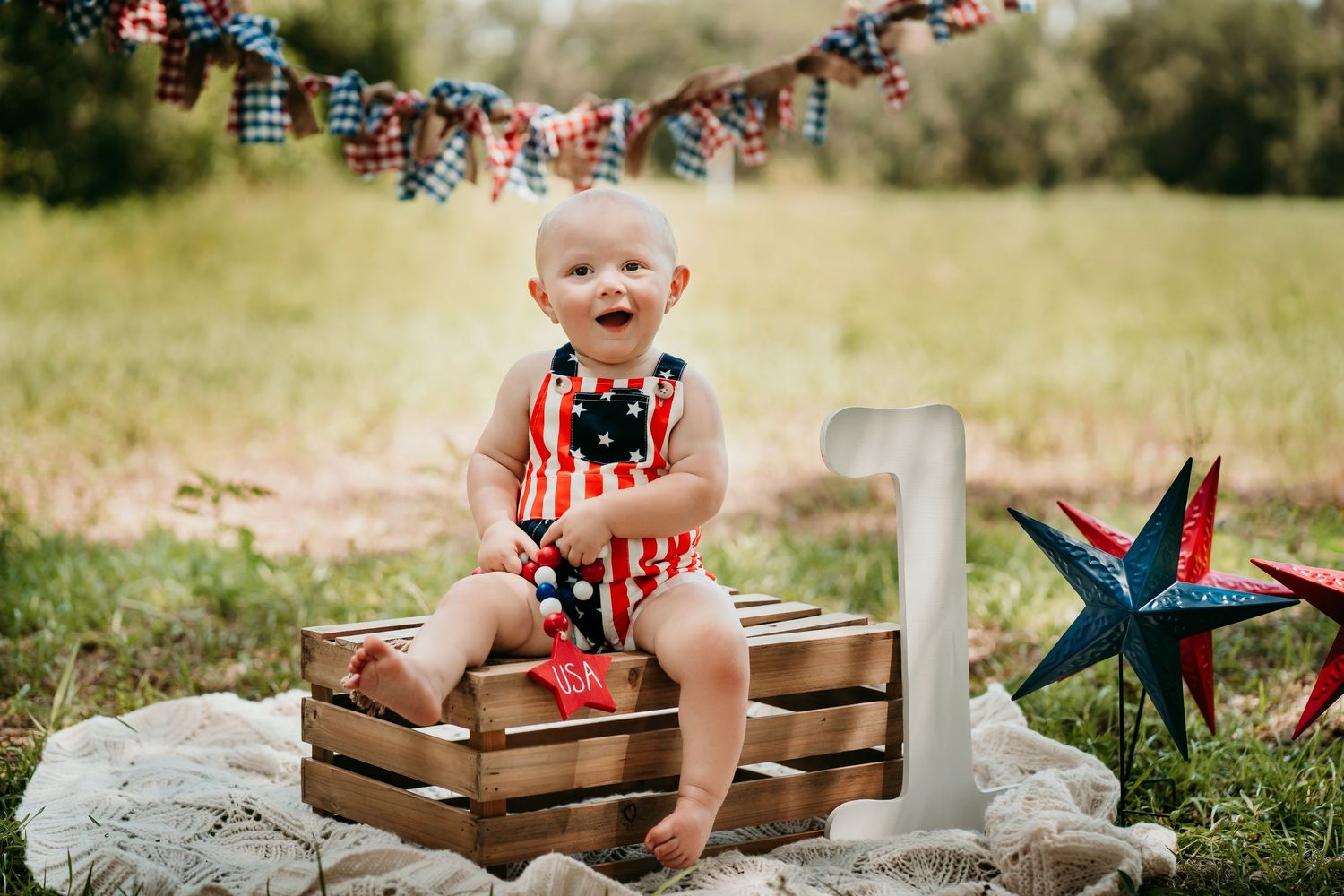 Baby's first birthday photo with patriotic decor, wooden crate, and number one sign outdoors.