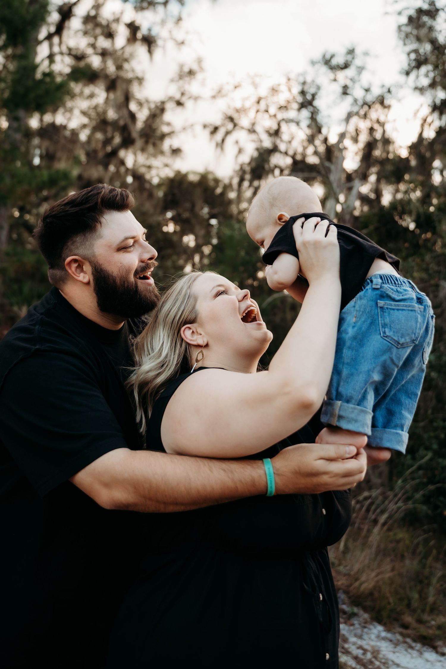 Young couple laughing joyfully while lifting their baby outdoors in a wooded setting.