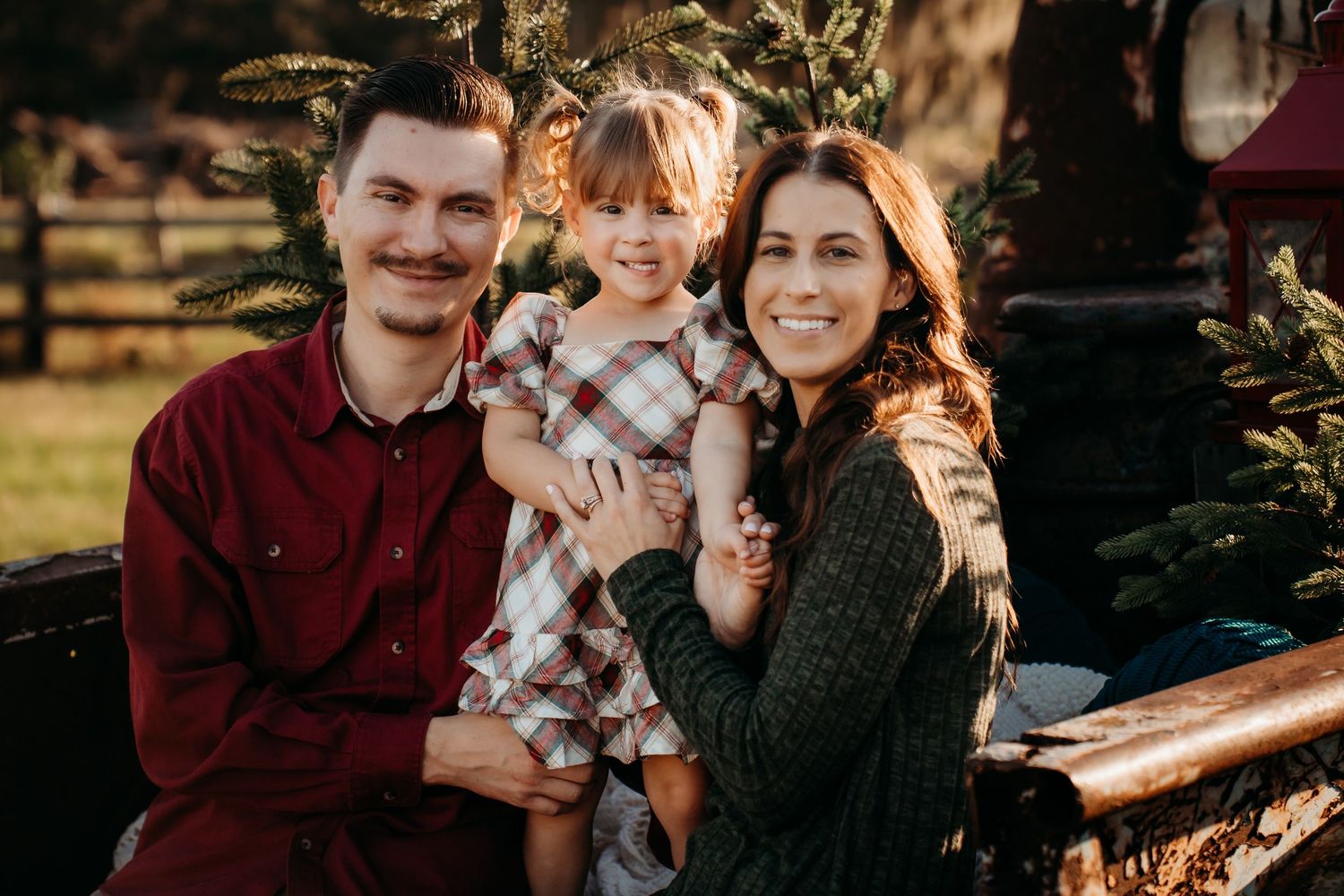 Happy family of three posing together at an outdoor Christmas tree farm during sunset.