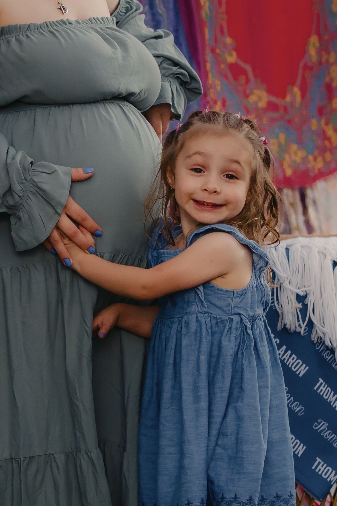 A smiling toddler girl in a blue dress hugs a pregnant woman's belly during a maternity photo session.