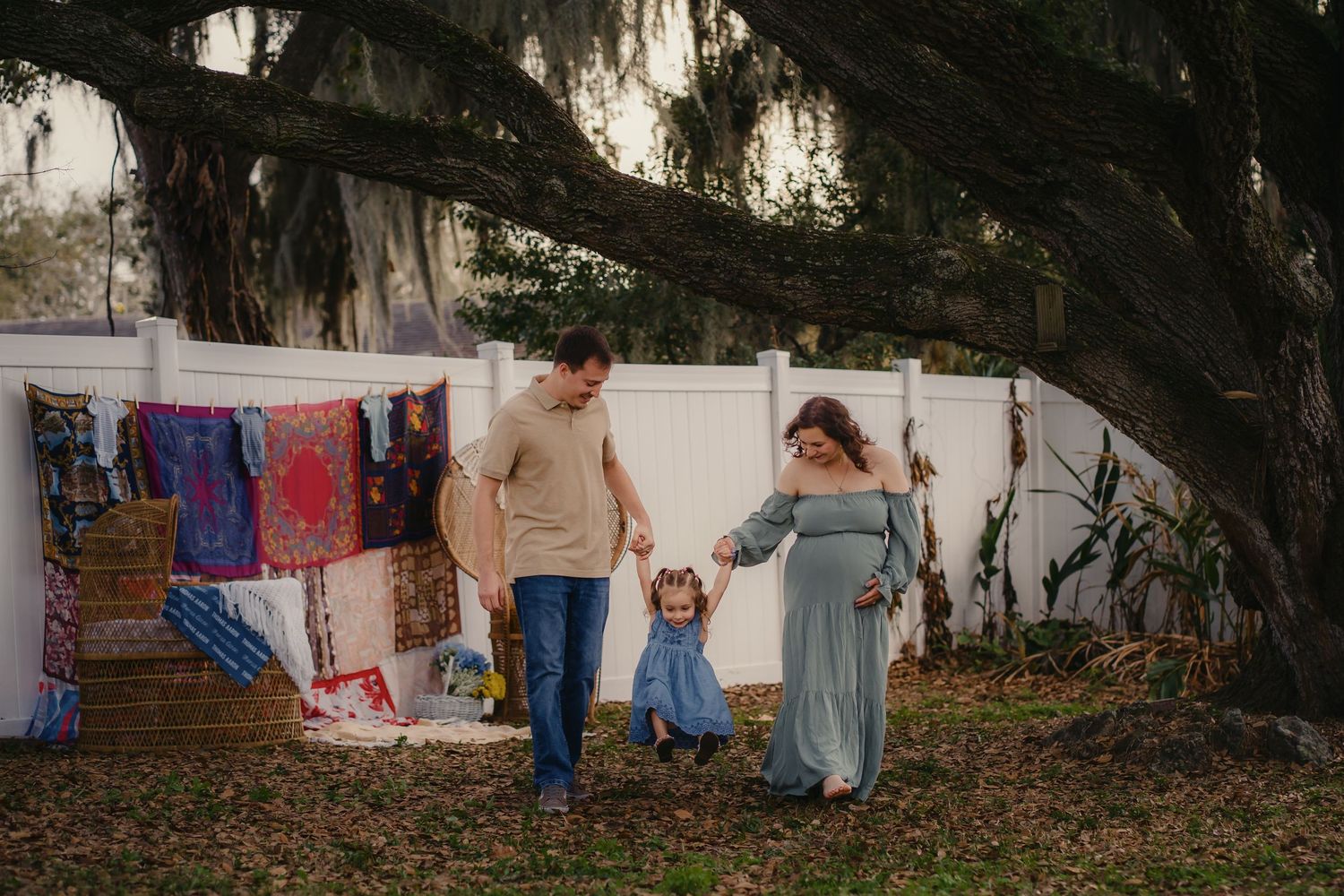 Pregnant woman in blue dress with husband and toddler daughter holding hands outdoors near colorful quilts and large oak tree.