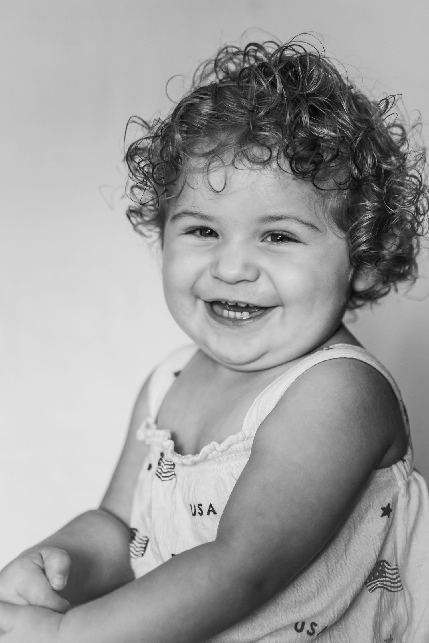 Black and white portrait of a happy toddler with curly hair wearing a USA dress, smiling brightly.