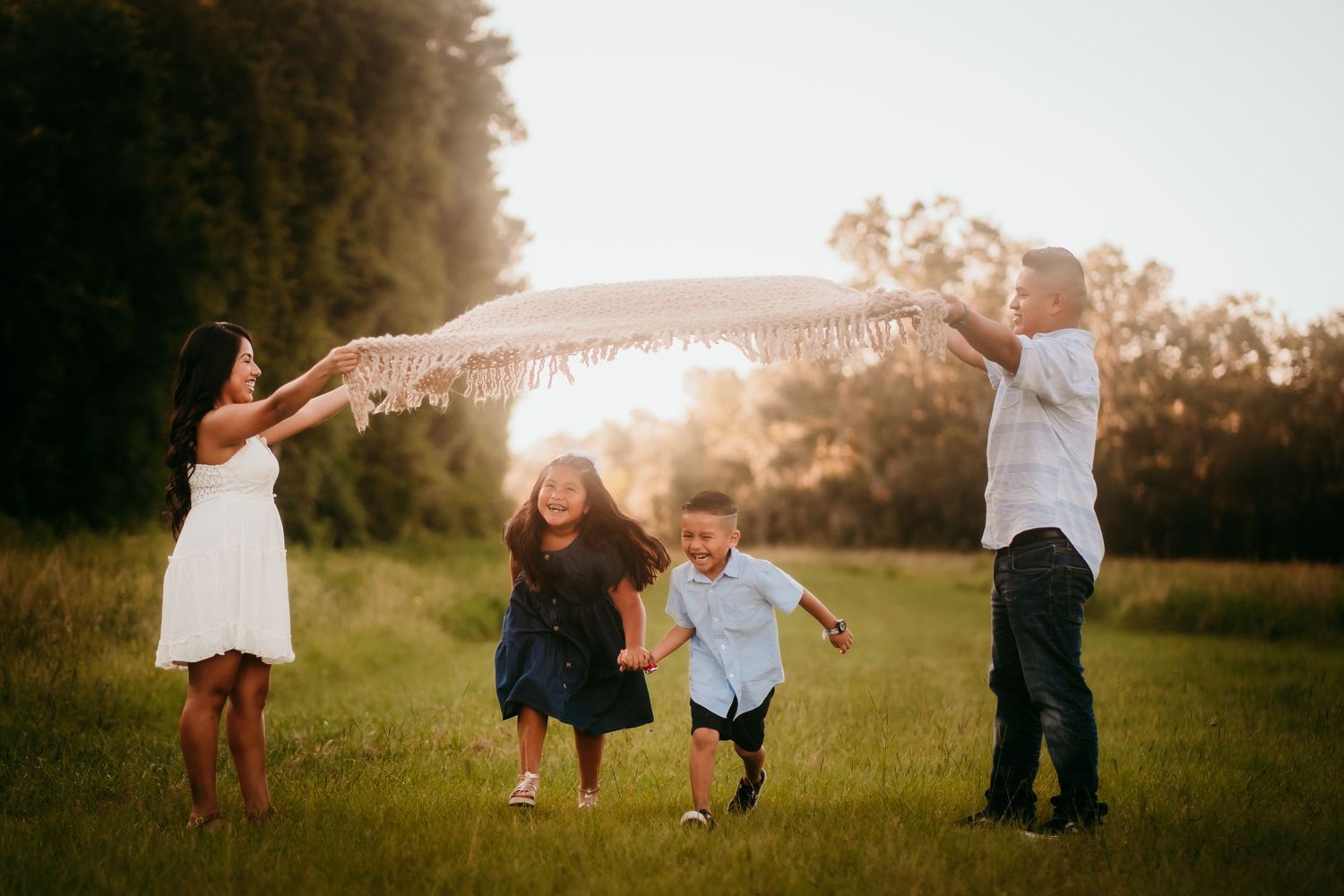 Joyful family of four playing under a blanket in a sunlit green field at sunset.