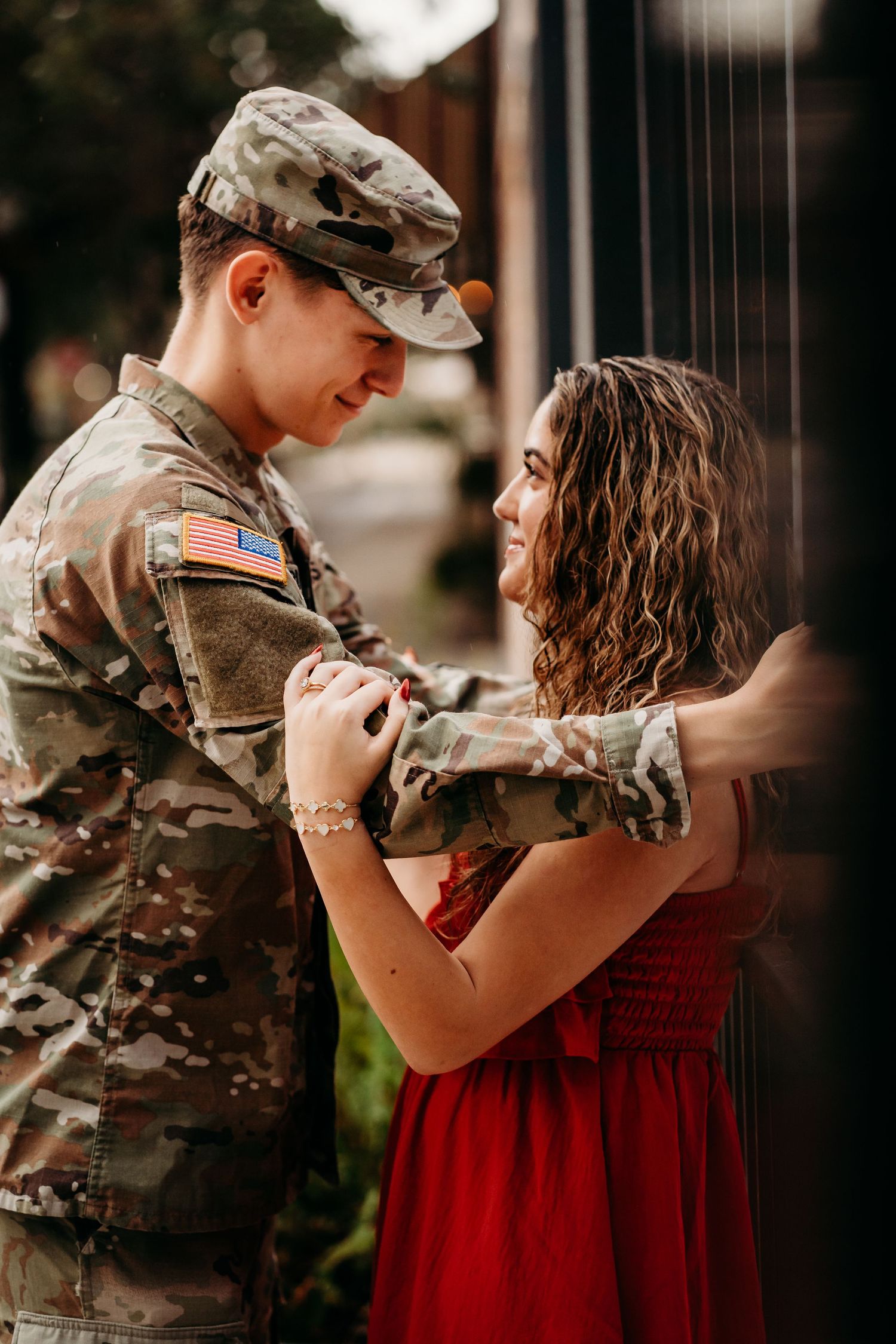 US Army soldier in camouflage uniform holding hands with smiling woman in red dress outdoors.