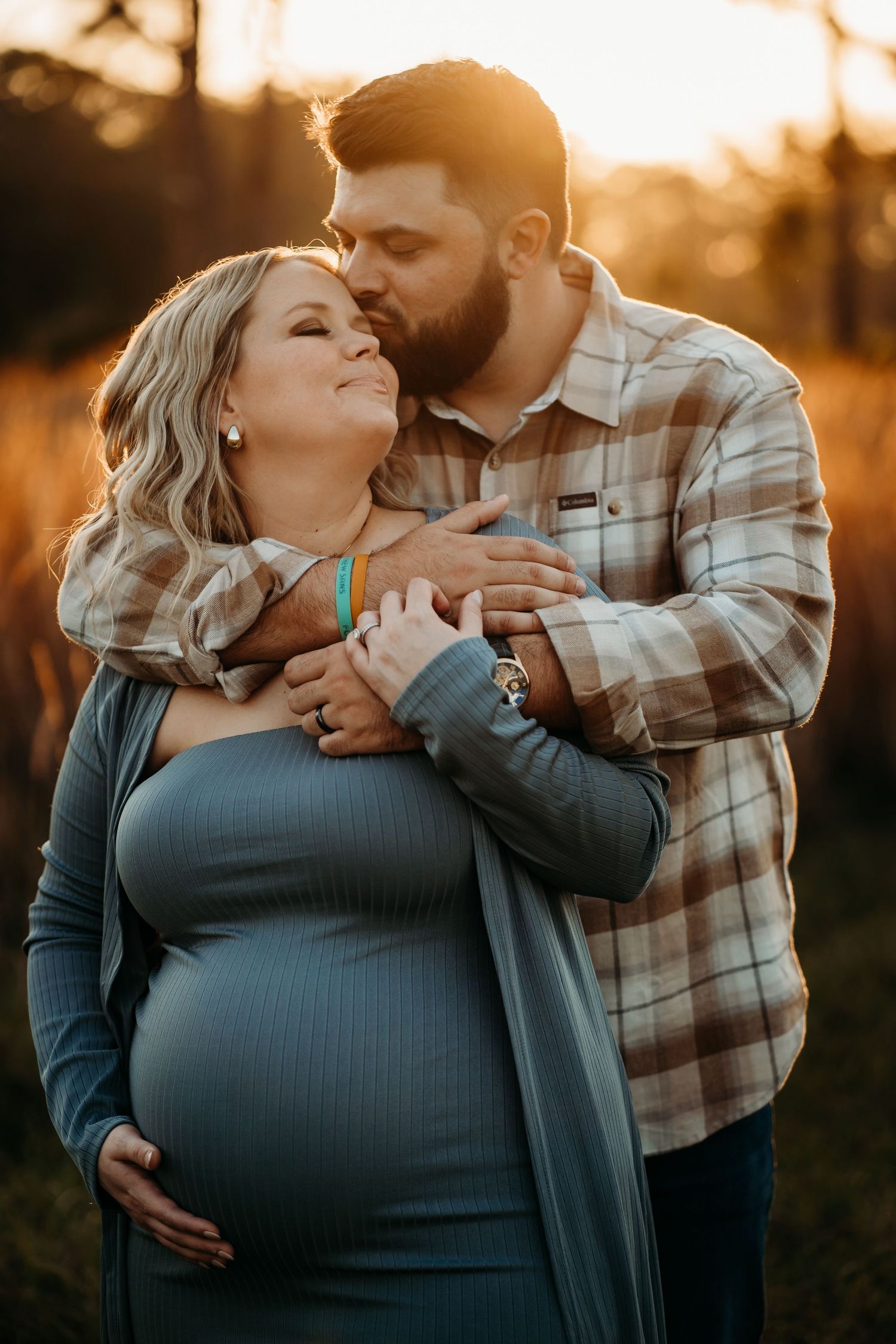Man kissing pregnant woman's cheek while embracing her from behind at golden hour outdoors.