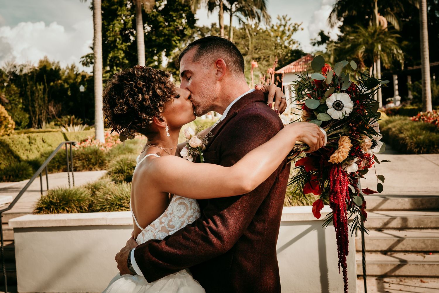 Bride and groom share a romantic kiss outdoors, with bride holding a lush burgundy and white wedding bouquet.