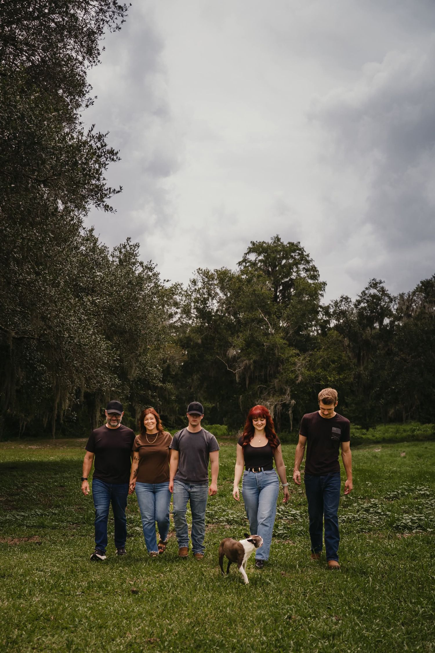 Five people and a dog pose in a green field surrounded by trees under a cloudy sky.