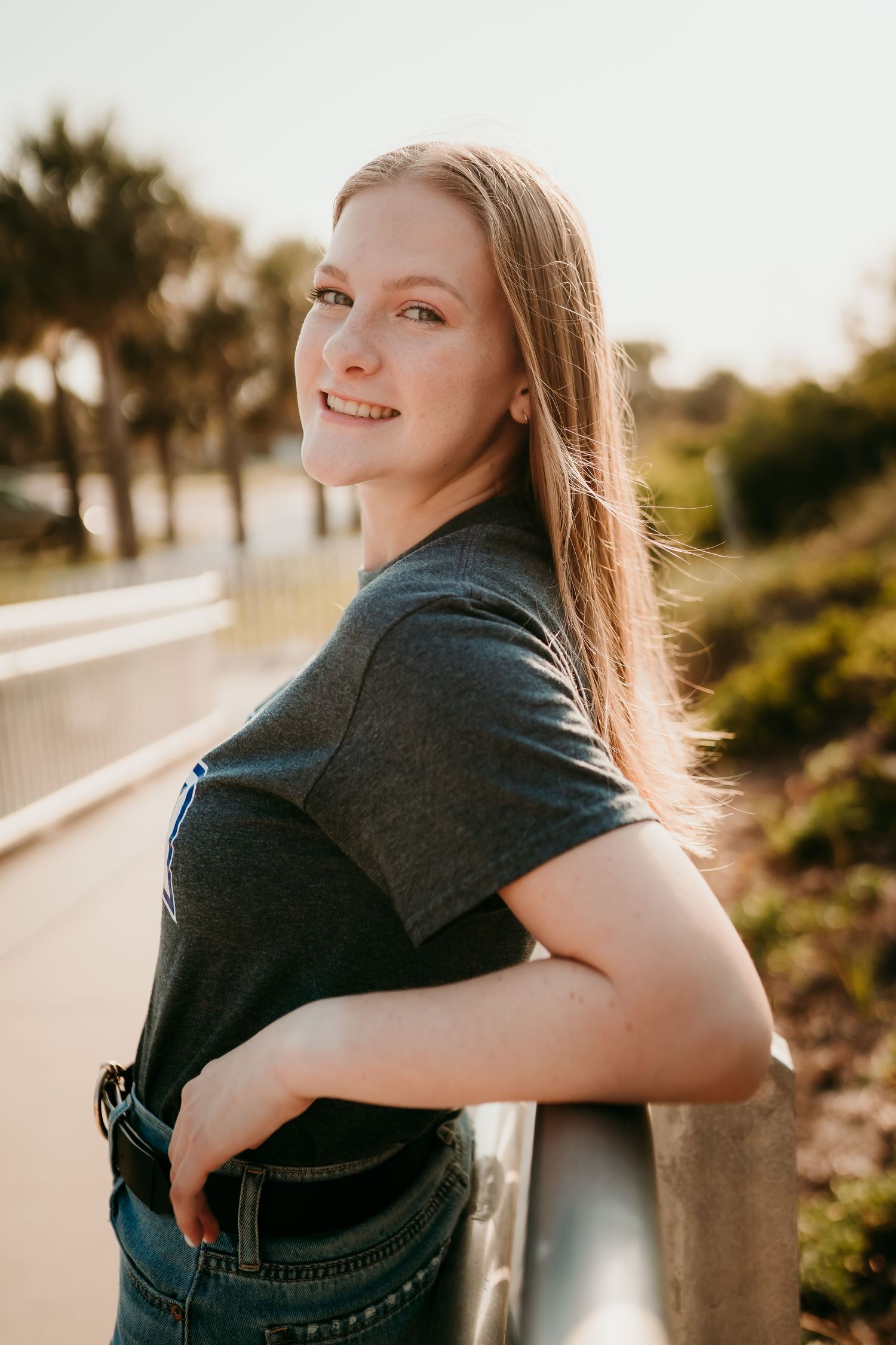 Young woman with long blonde hair smiling over her shoulder, leaning on a railing outdoors at golden hour.