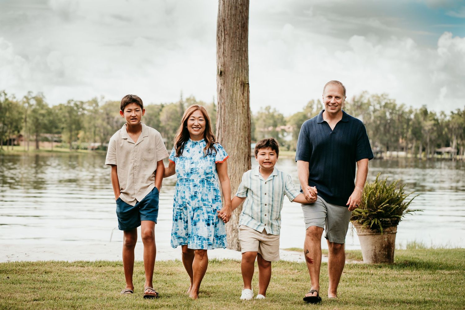 Smiling family of four walking hand-in-hand on green grass near a lakeside.