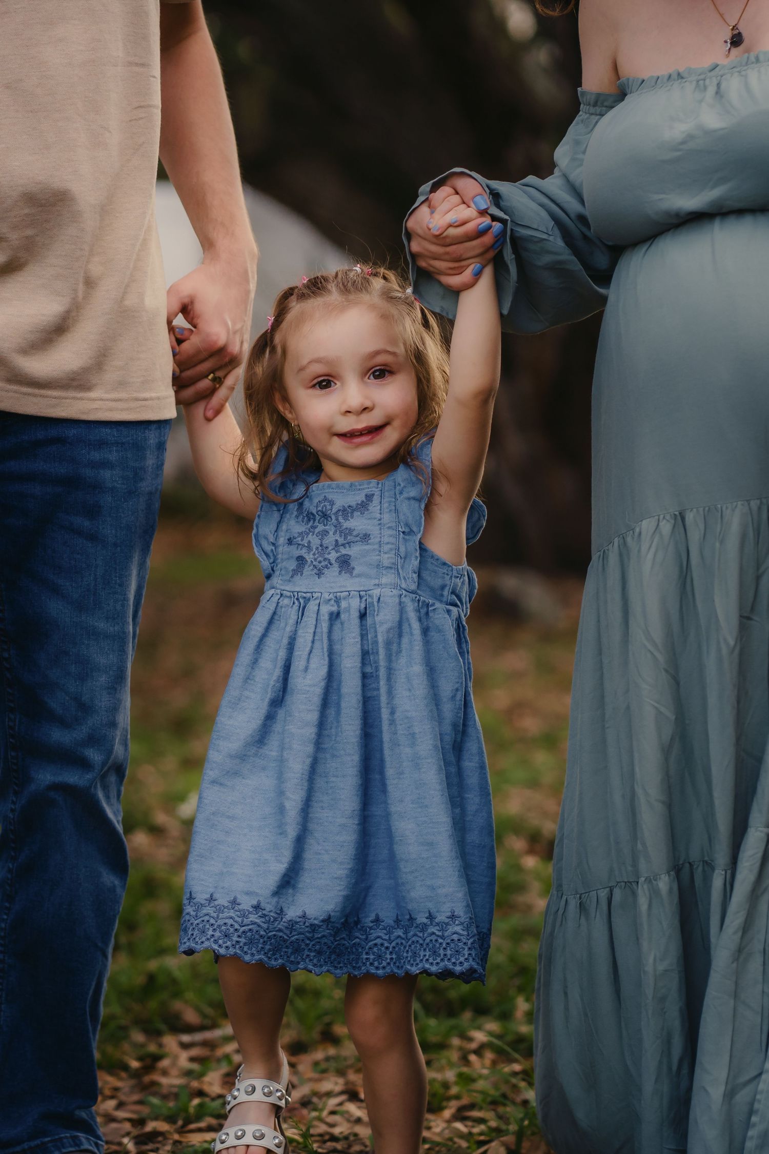 A smiling toddler girl in a blue dress holds hands with her parents outdoors in a sweet family portrait.