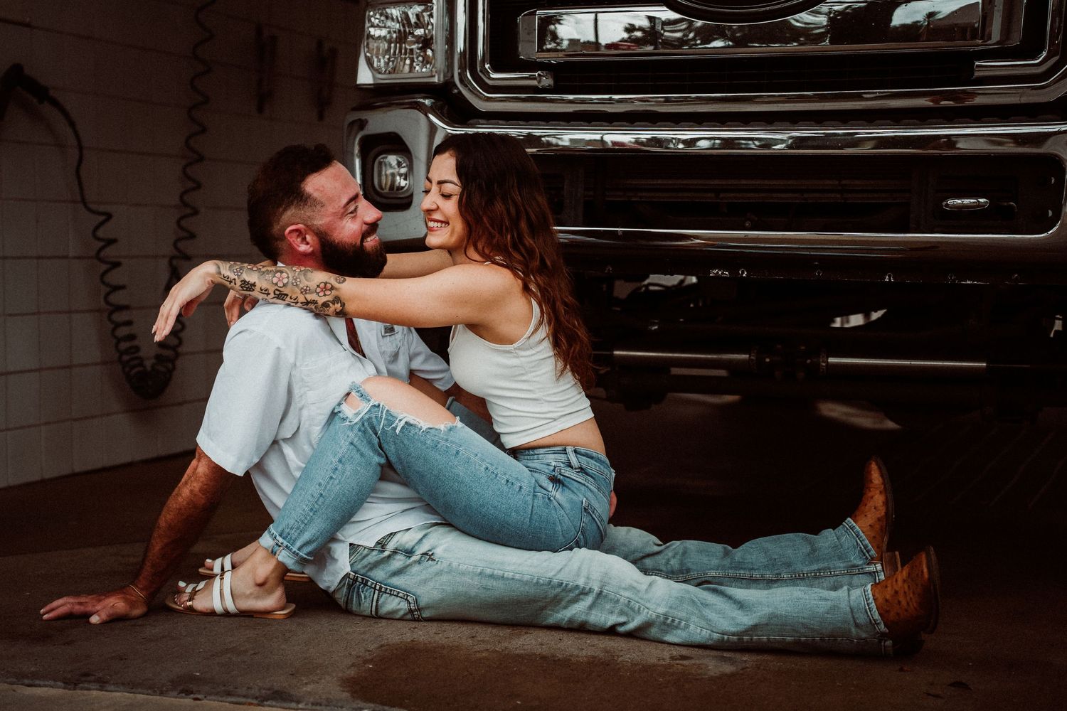 Smiling couple in jeans sitting together on garage floor beside classic truck.
