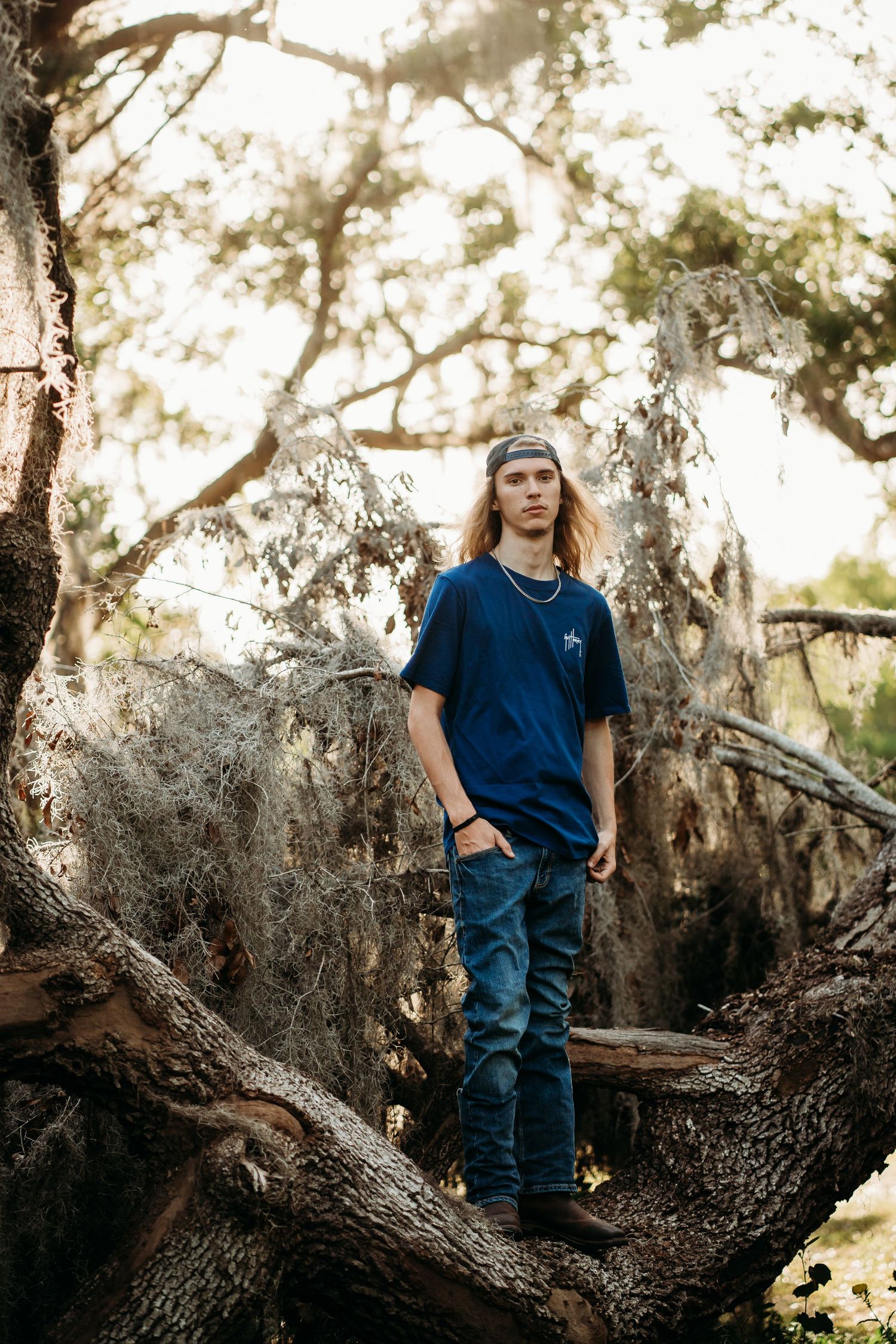 Young man with long hair in blue t-shirt and jeans standing on tree roots in a sunlit forest.