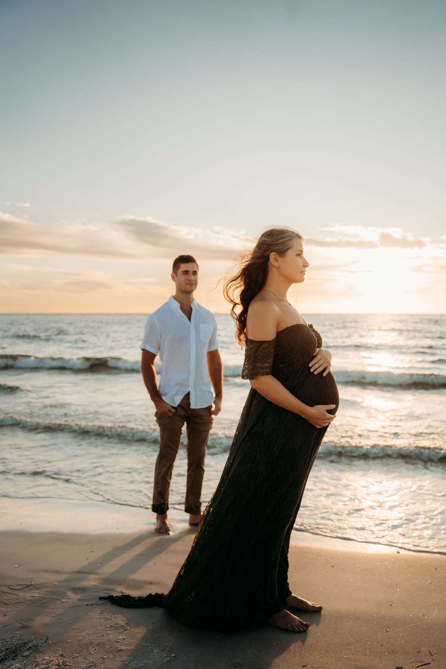 A pregnant woman in a black lace gown stands on a rocky beach at sunset while her partner watches from behind.