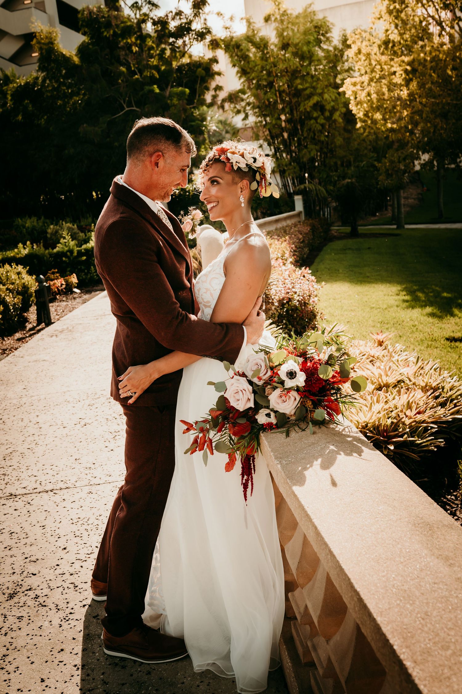 Bride in white gown with floral crown holds bouquet while embracing groom in brown suit outdoors on sunny day.