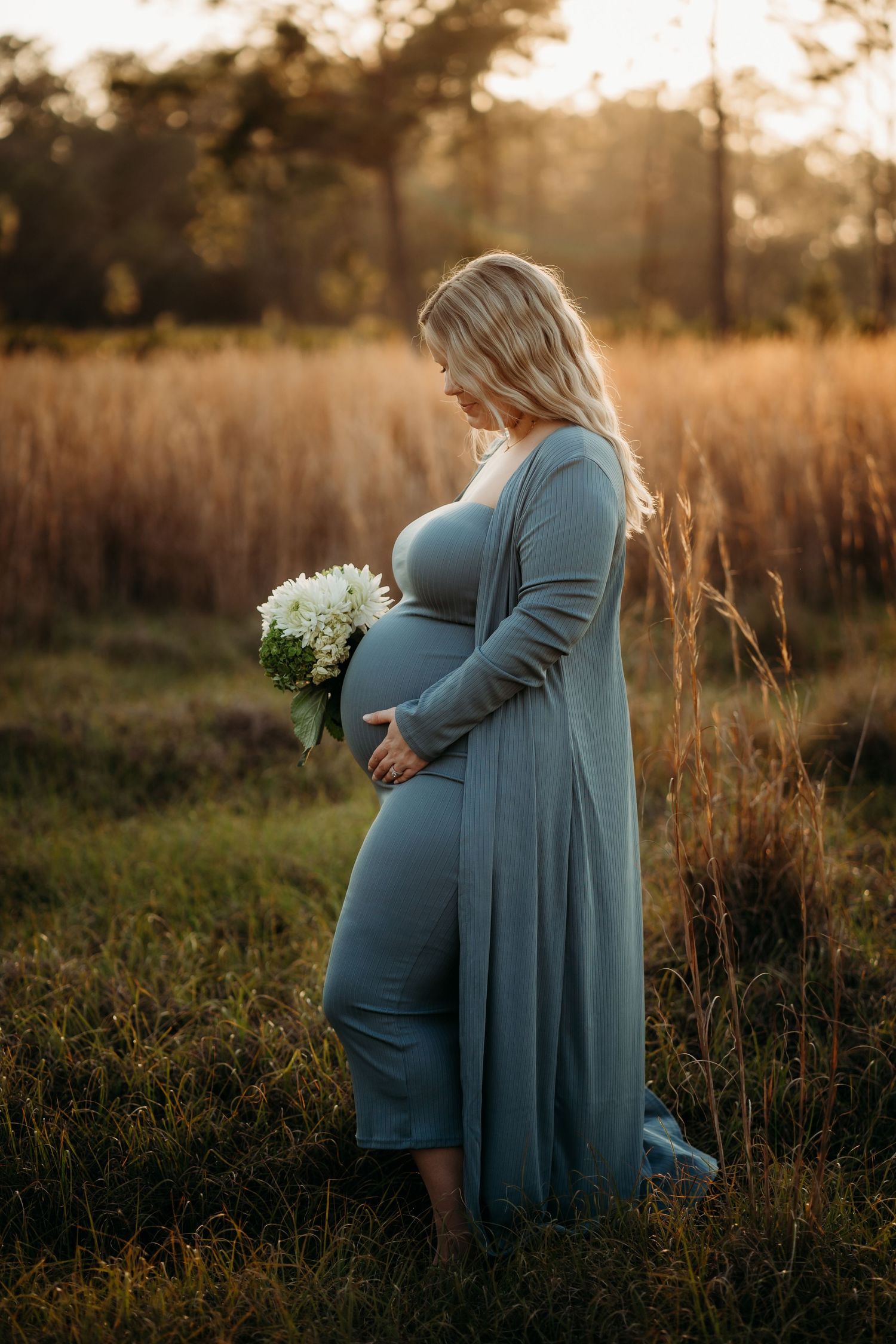 Pregnant woman in teal maxi dress holding white flowers at golden hour in a field.