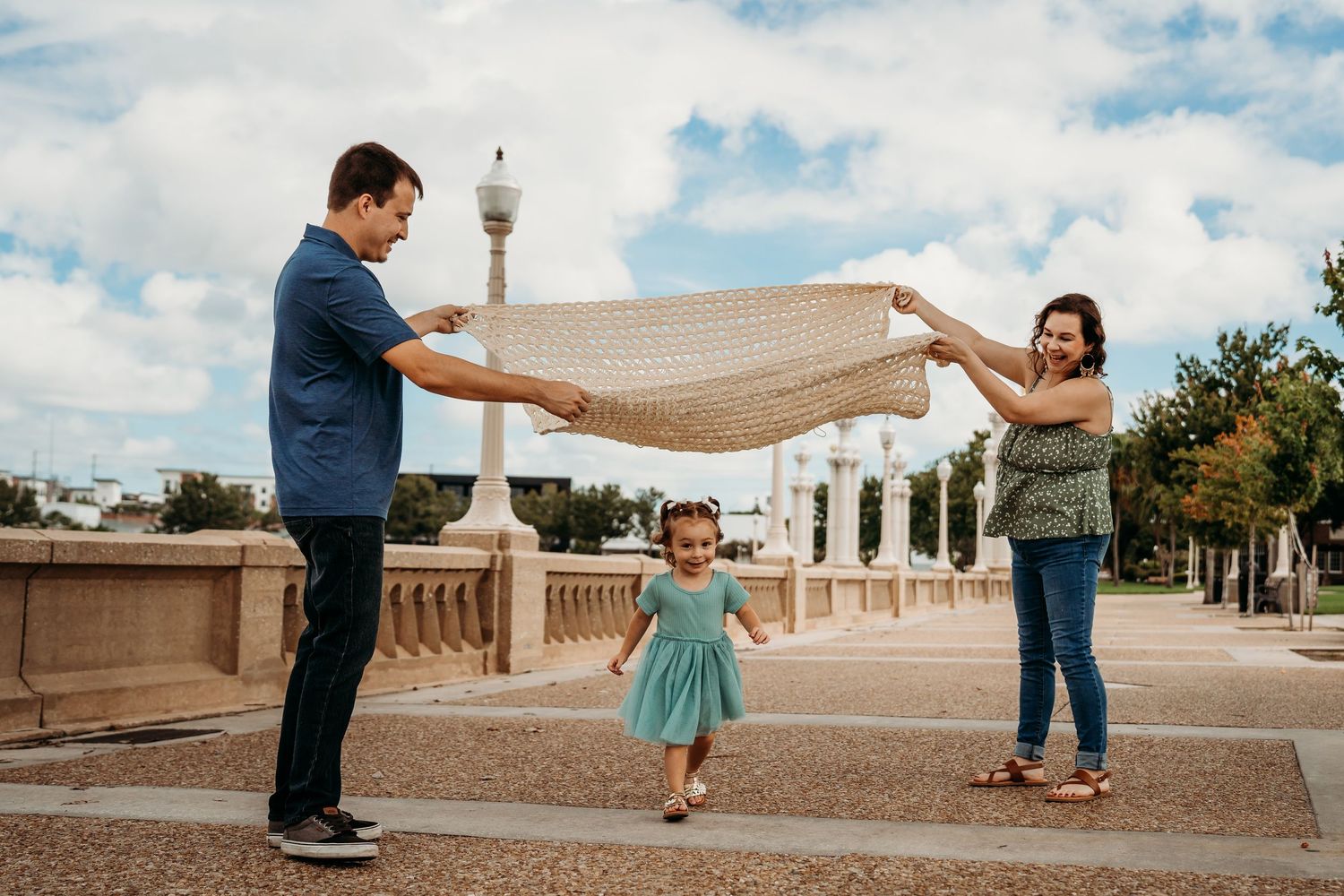 Parents holding a blanket over their toddler daughter walking on a sunny plaza.