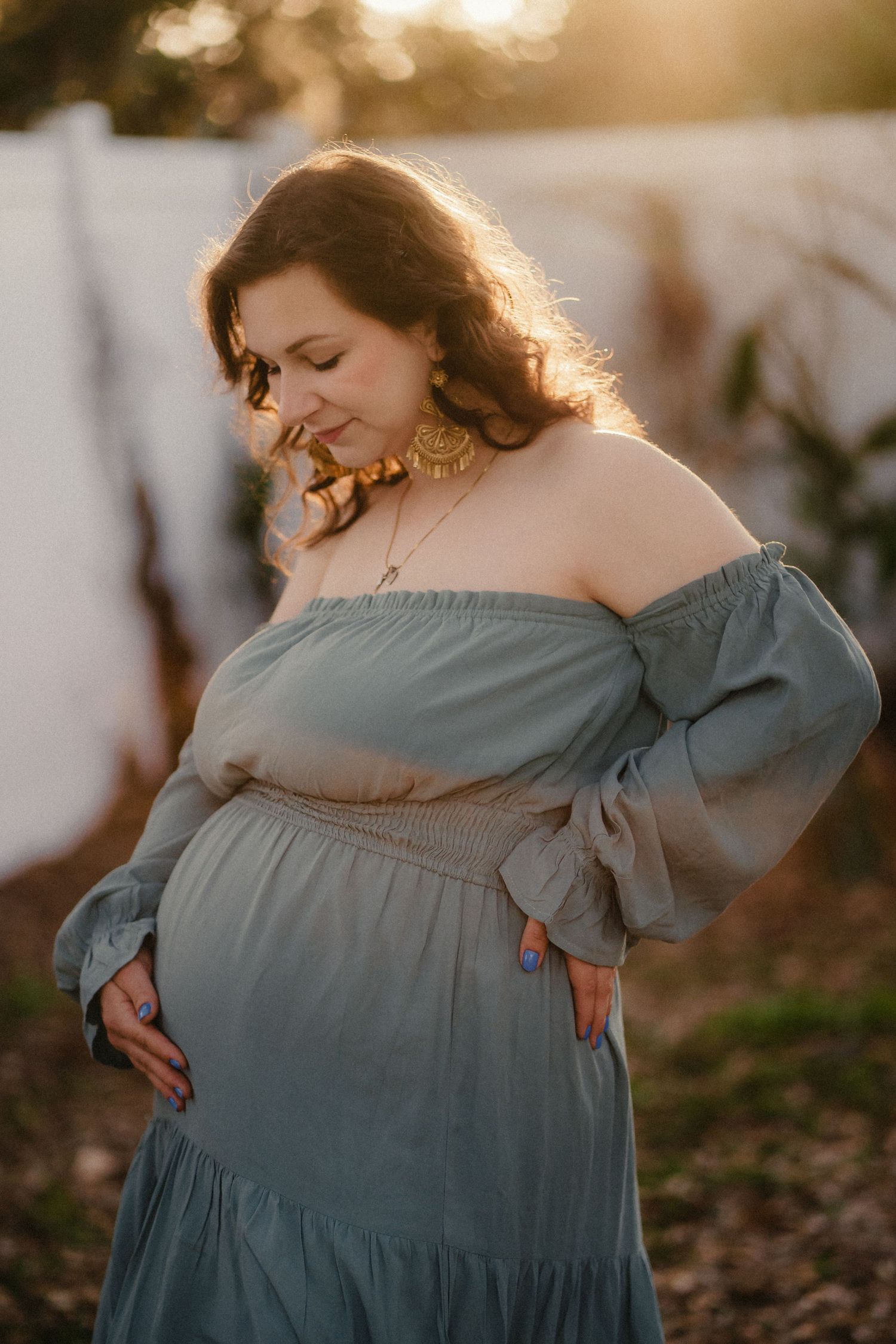 Pregnant woman in off-shoulder sage green dress with gold earrings, posing outdoors in golden hour sunlight.