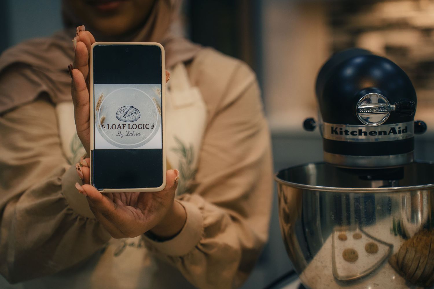 Person holding smartphone displaying Loaf Logic logo beside a KitchenAid stand mixer.