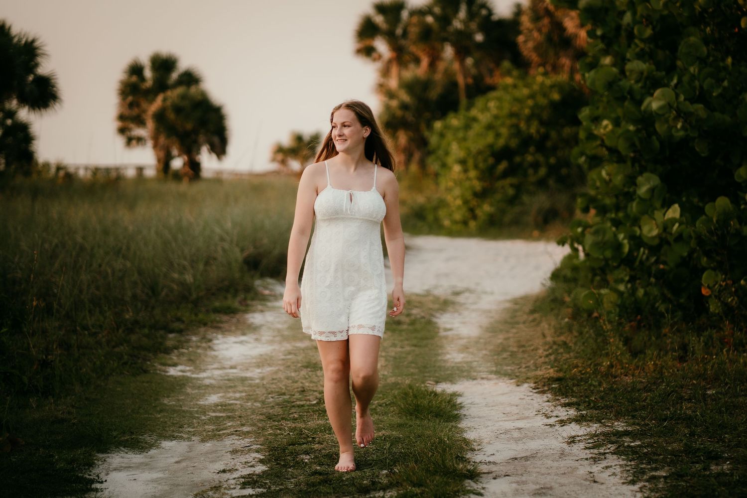 Young woman in white lace dress walking barefoot on sandy path surrounded by lush green trees at sunset.