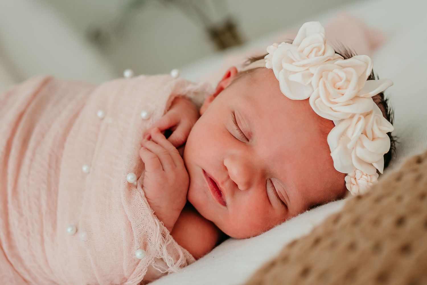 Sleeping newborn baby girl wrapped in pink with pearl details and floral headband.