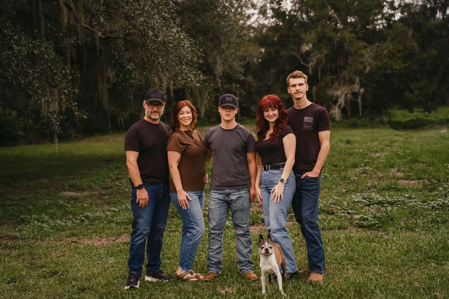 Family of five posing outdoors in matching dark shirts and jeans with a small dog on a green grassy field.