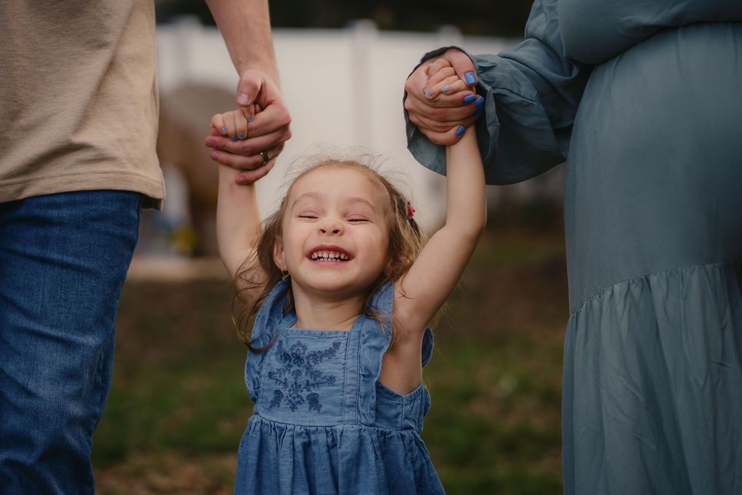 Happy toddler girl in blue dress holds parents hands outdoors, smiling with eyes closed in joyful family moment.