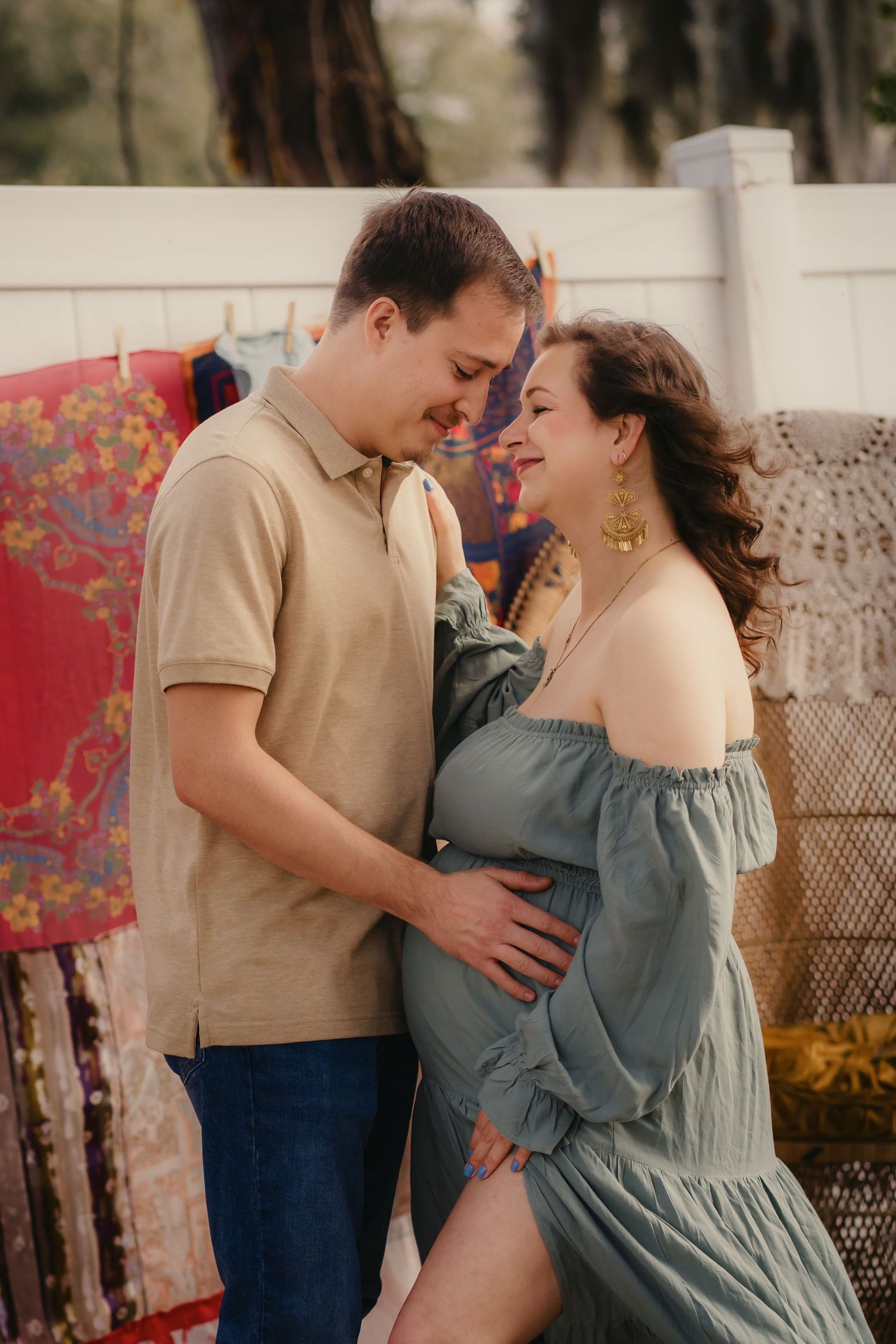 Couple sharing a tender moment outdoors, woman in sage off-shoulder maternity dress, man in tan polo, colorful fabrics behind them.