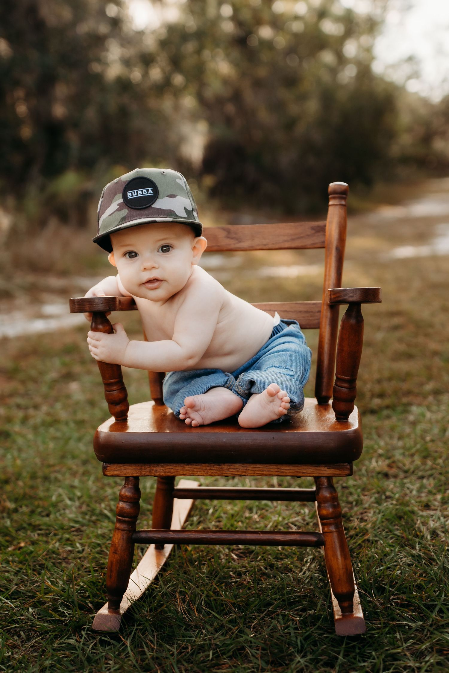 Shirtless baby in camo hat and denim shorts sitting in a wooden rocking chair outdoors on green grass.