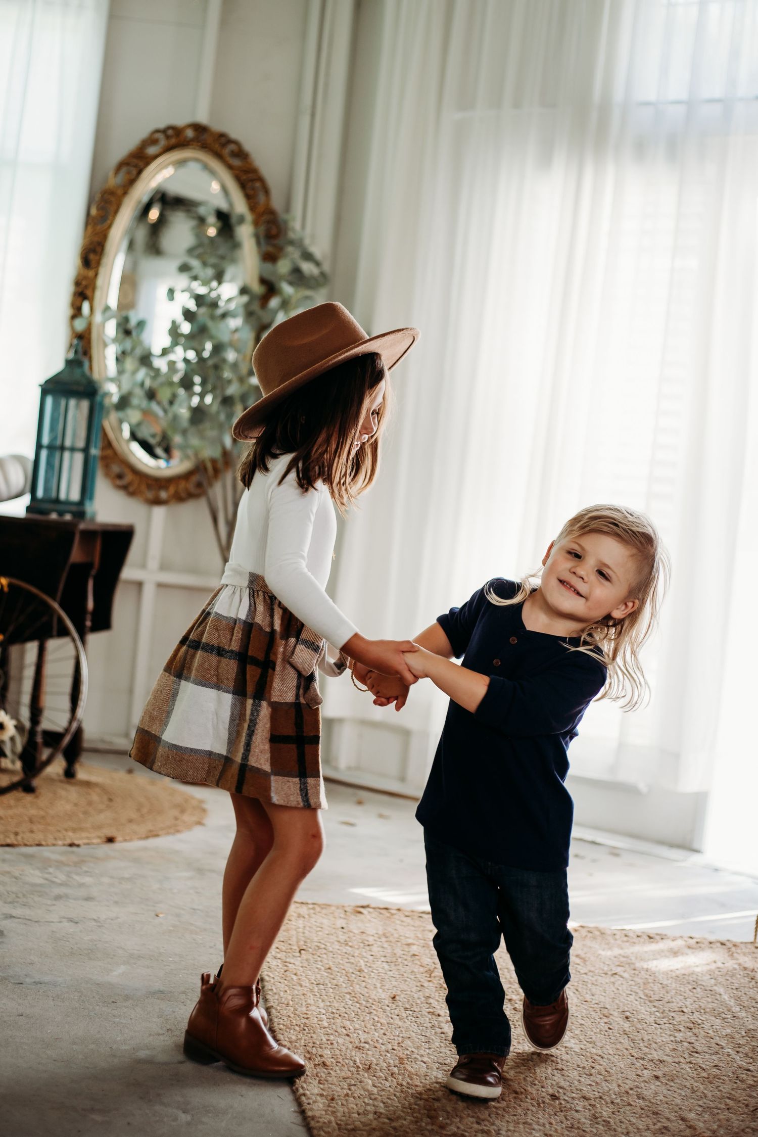 Two stylish children holding hands and dancing indoors, girl wearing plaid skirt and brown hat.