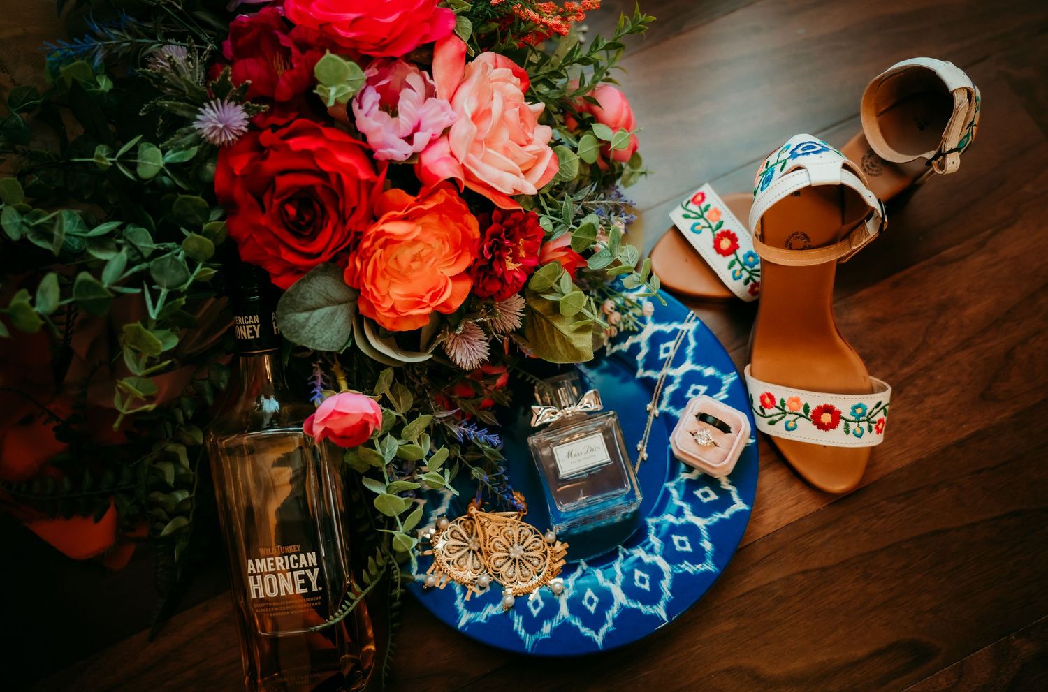 Flat lay of red roses, American Honey bottle, perfume, floral sandals, and blue tray on wooden surface.