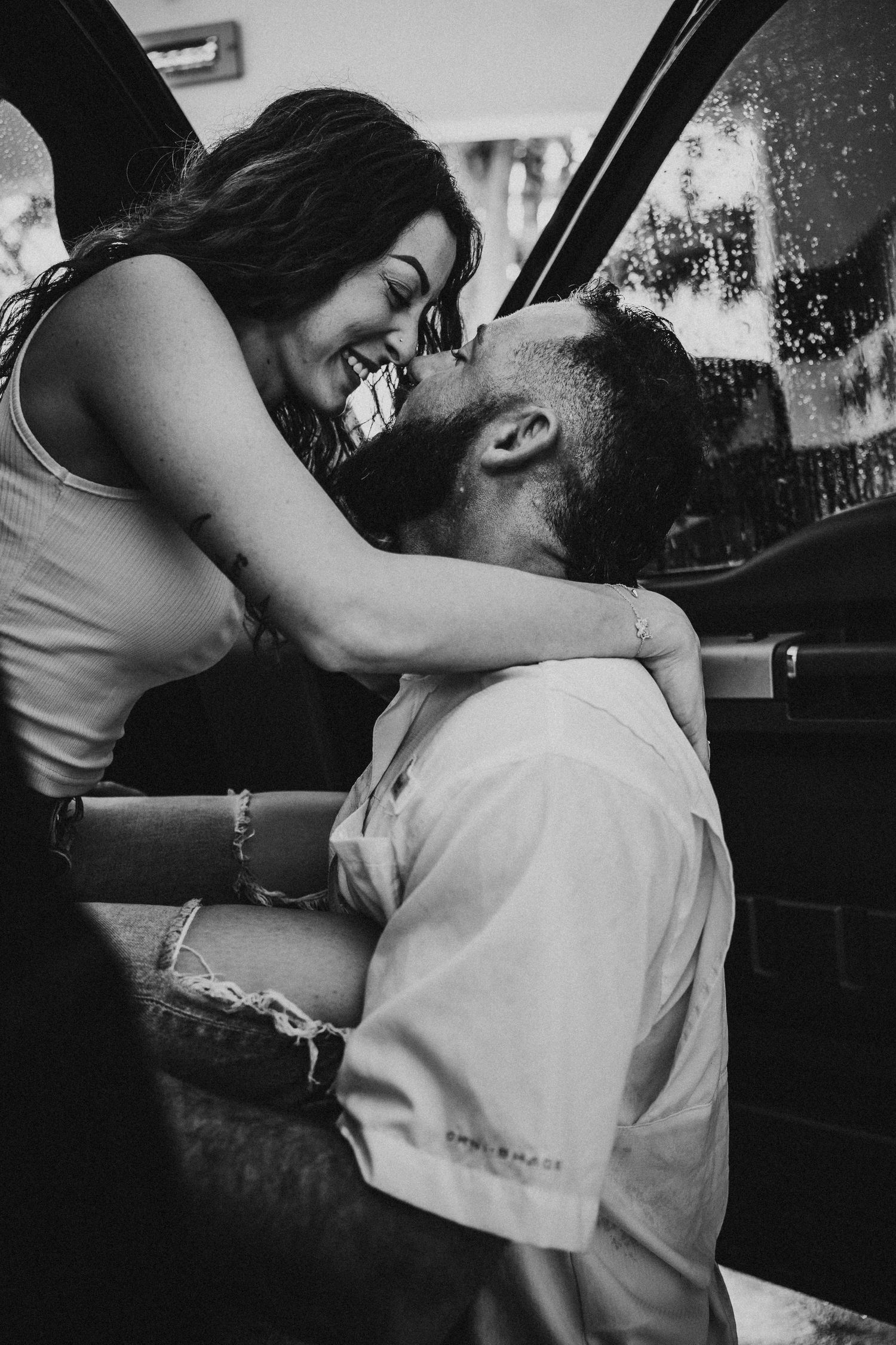 Black and white photo of smiling couple sharing a kiss beside a vehicle window.