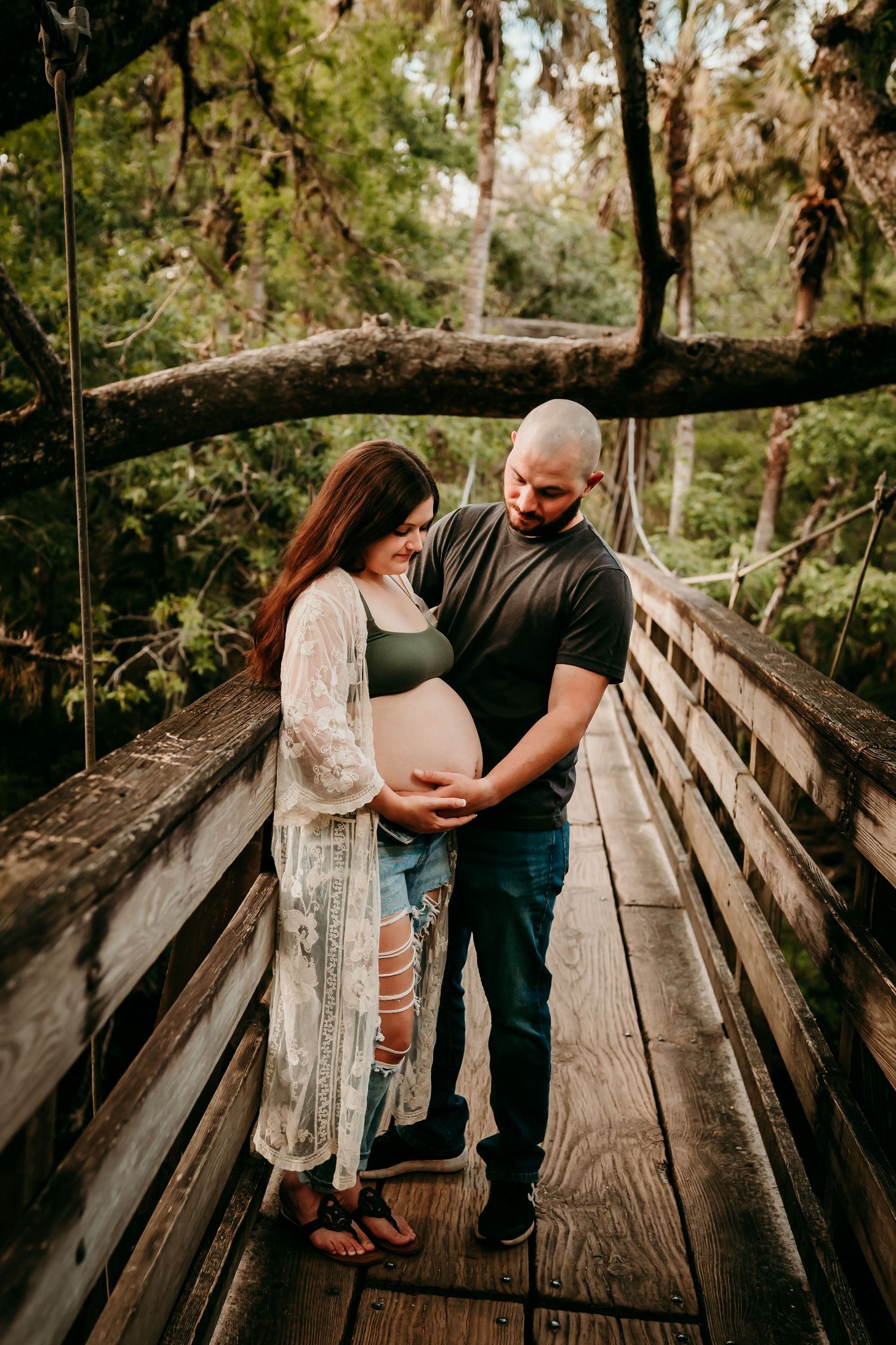 Expecting couple holding baby bump together on a rustic wooden bridge surrounded by trees.