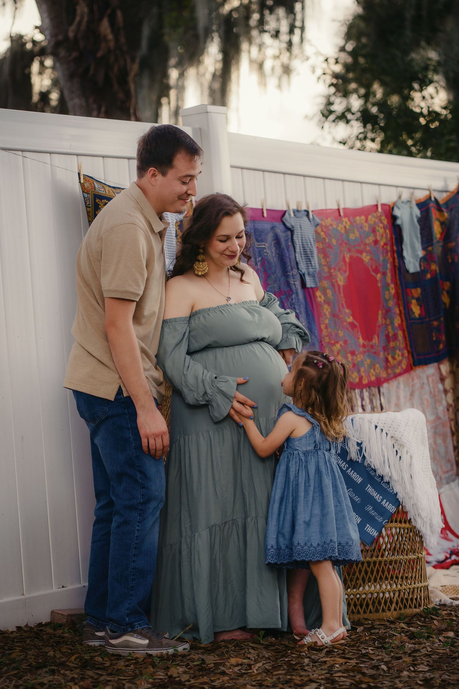 Pregnant woman in sage green dress with husband and young daughter touching baby bump during outdoor maternity photo session.