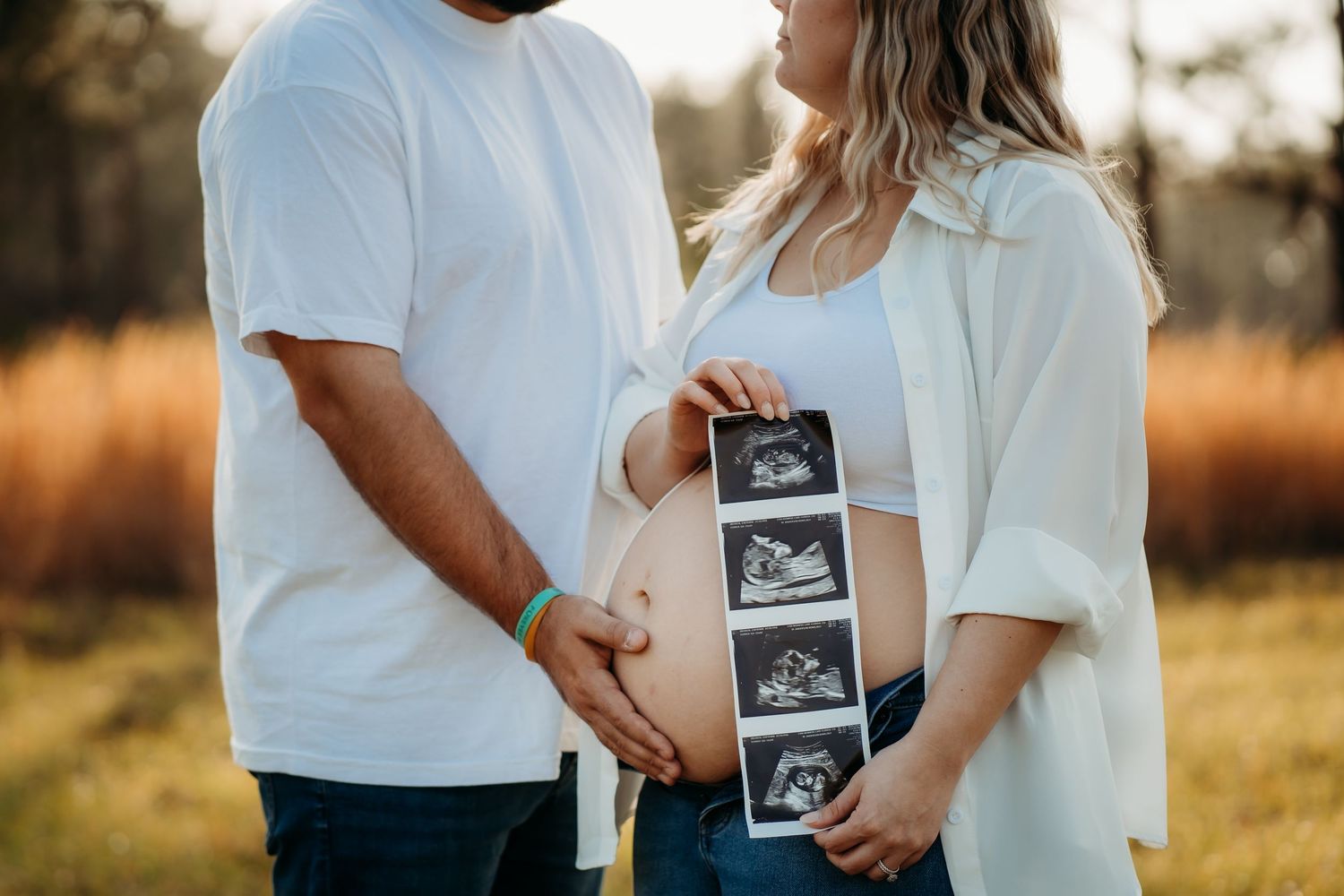 Couple holding ultrasound photos in front of pregnant belly during outdoor maternity session.