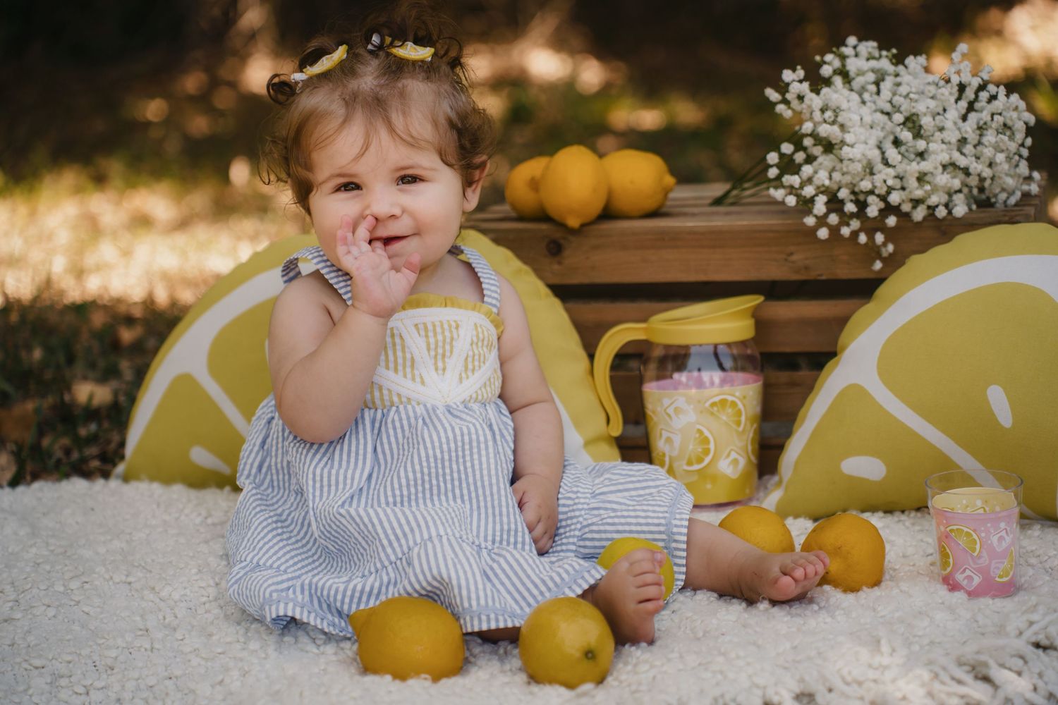 Adorable toddler girl in striped dress sitting among lemons with lemon slice decorations for a sweet photo shoot.