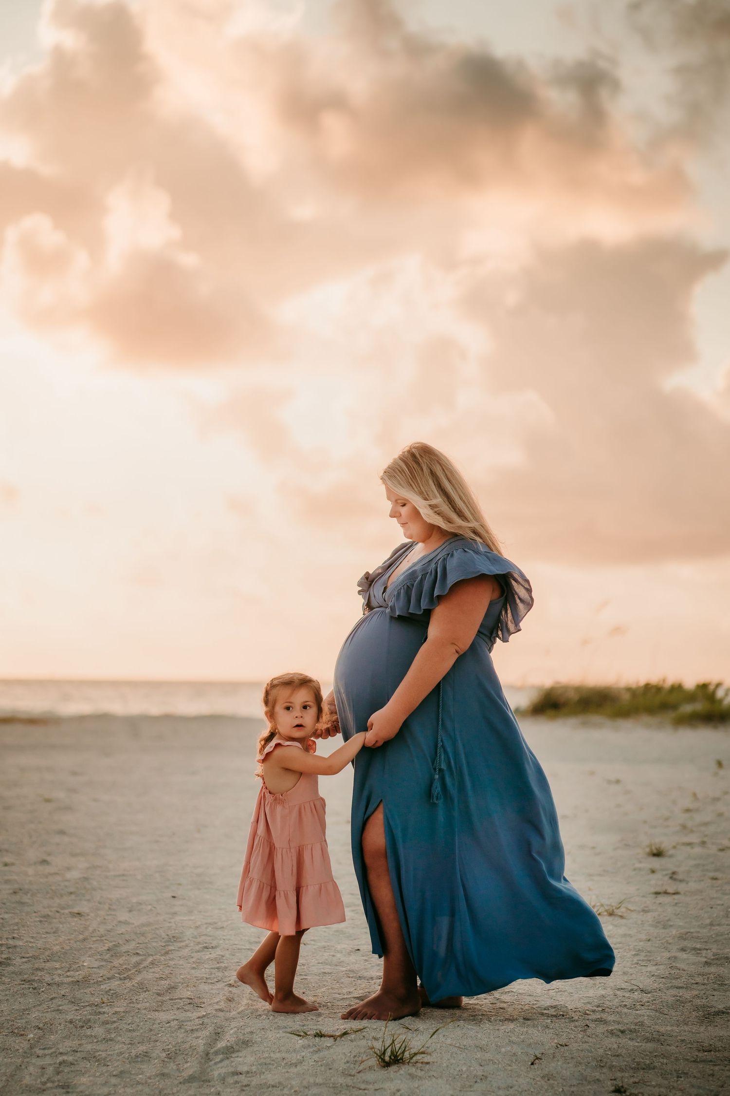 Pregnant woman in blue dress holding hands with toddler daughter on sandy beach at sunset.